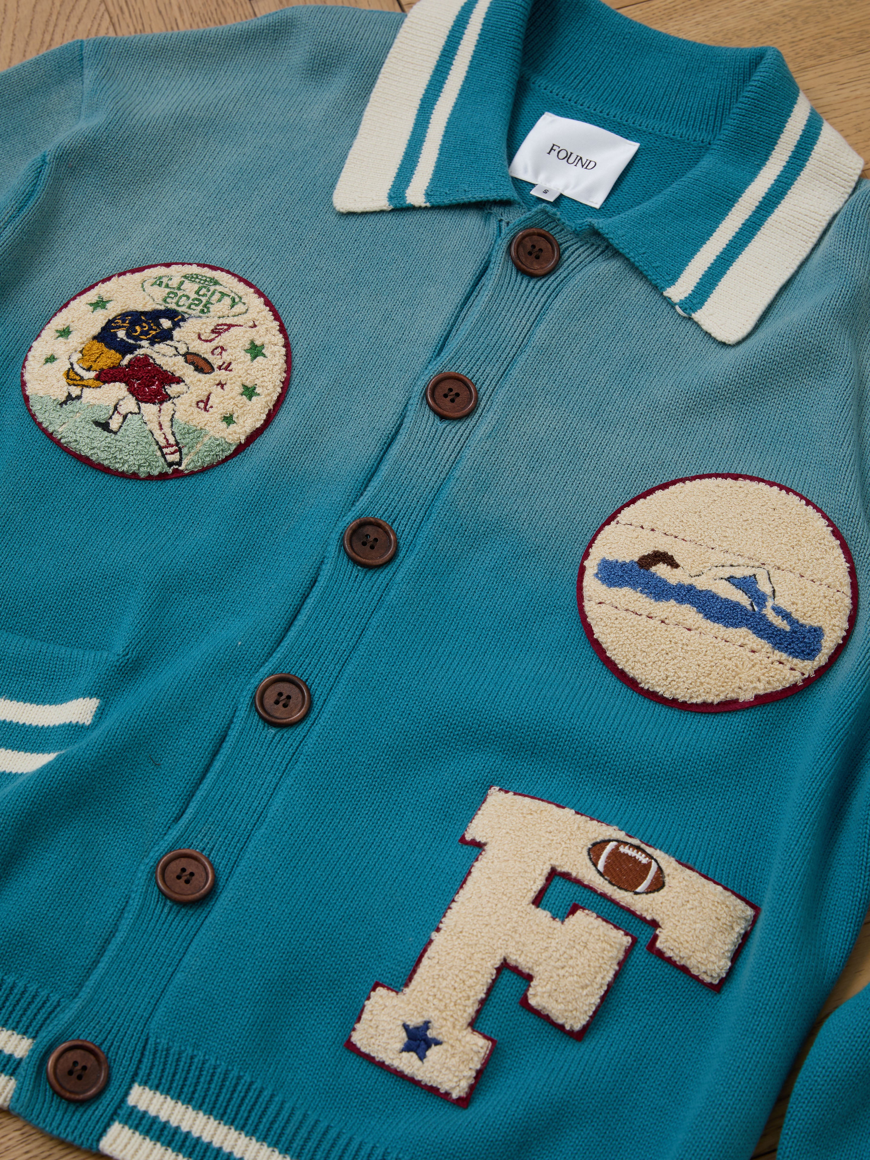 The Fin Varsity Patch Collared Cardigan by FOUND features a teal design with brown buttons, sports-themed patches: a football scene, swimmer, and large "F" with football motif—ideal for those who love vintage collegiate style.