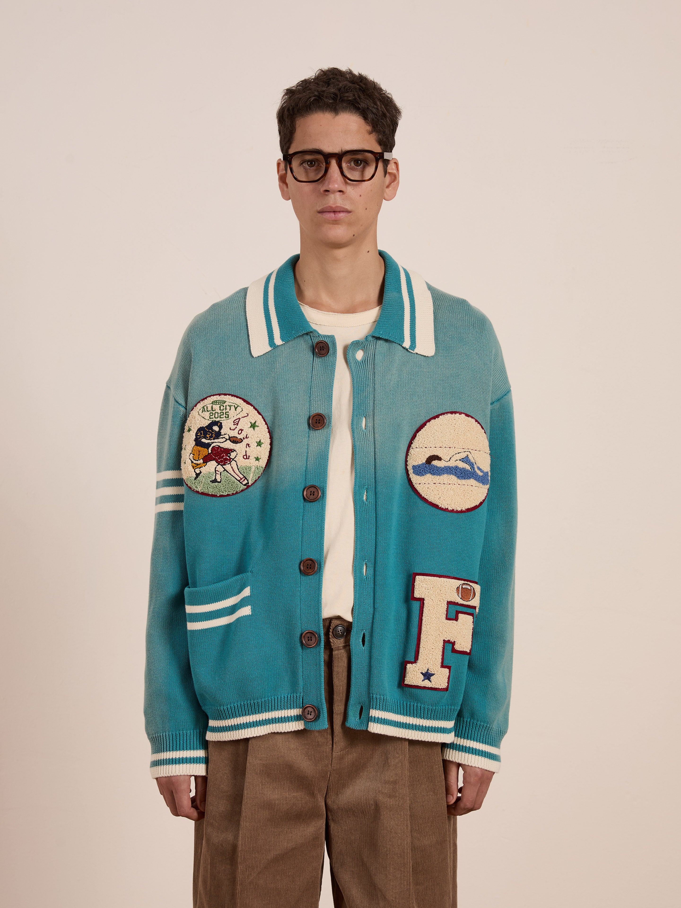 A person in glasses wears the Fin Varsity Patch Collared Cardigan by FOUND, featuring a vintage turquoise design, paired with brown pants. They stand facing forward against a plain background, exuding collegiate style.