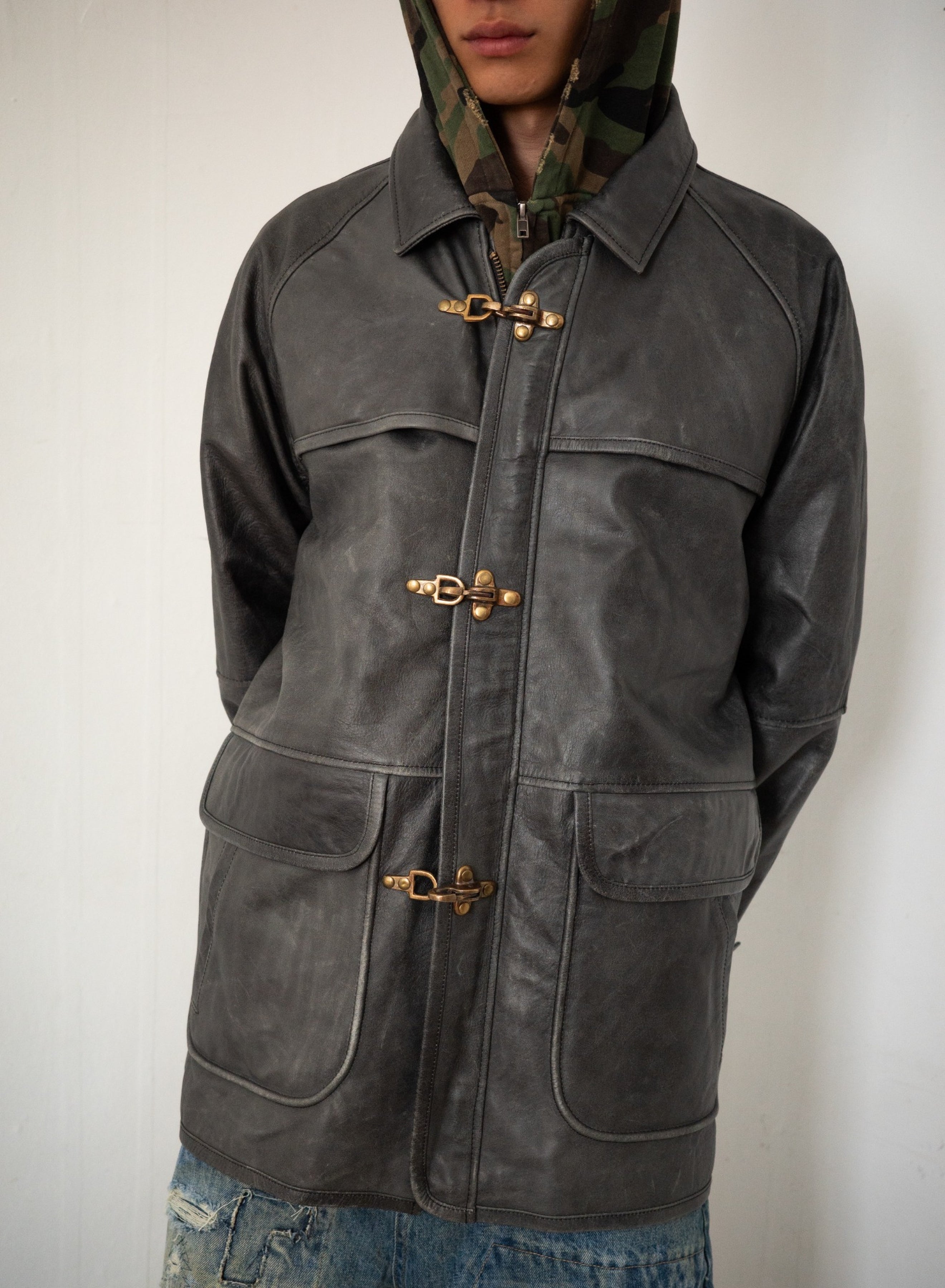 Model wears the FOUND Distressed Leather Fireman Clasp Jacket over a camouflage hoodie, hands behind their back, paired with blue distressed jeans for a unisex relaxed fit.