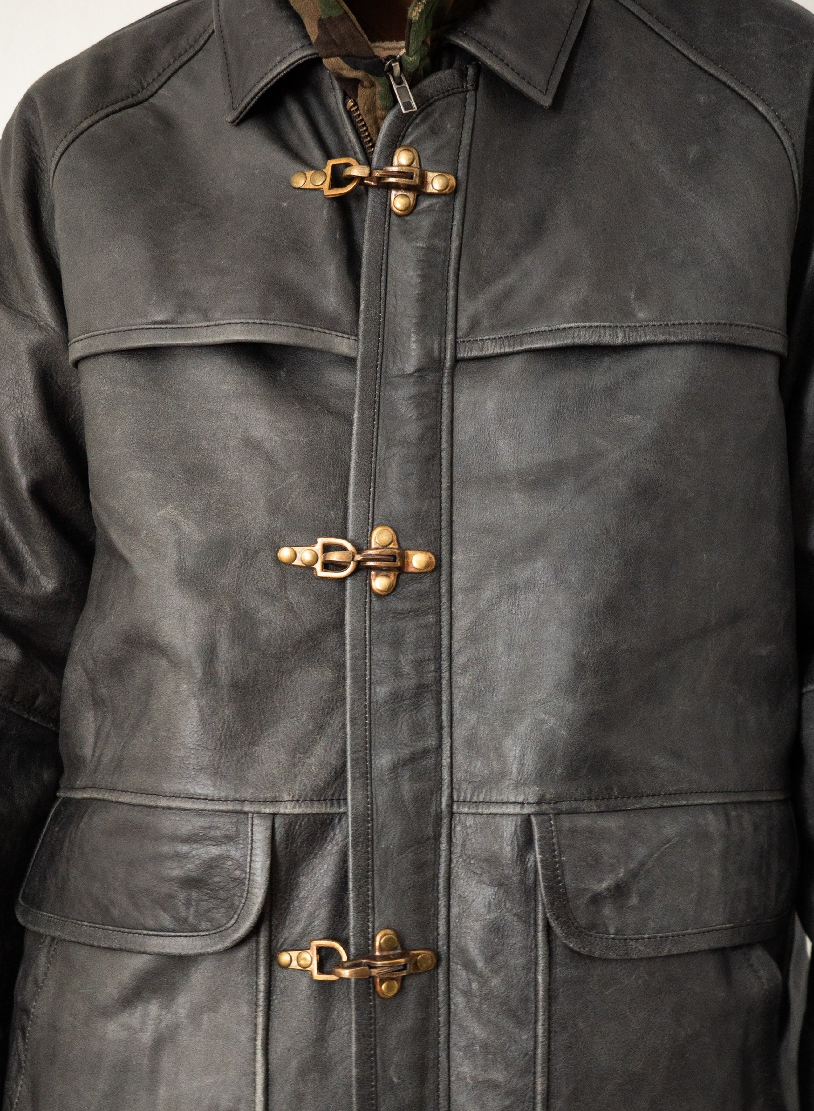 FOUND Distressed Leather Fireman Clasp Jacket in black leather features brass toggle clasps and two front pockets, worn over a camo shirt. This unisex jacket offers a relaxed fit for versatile style.