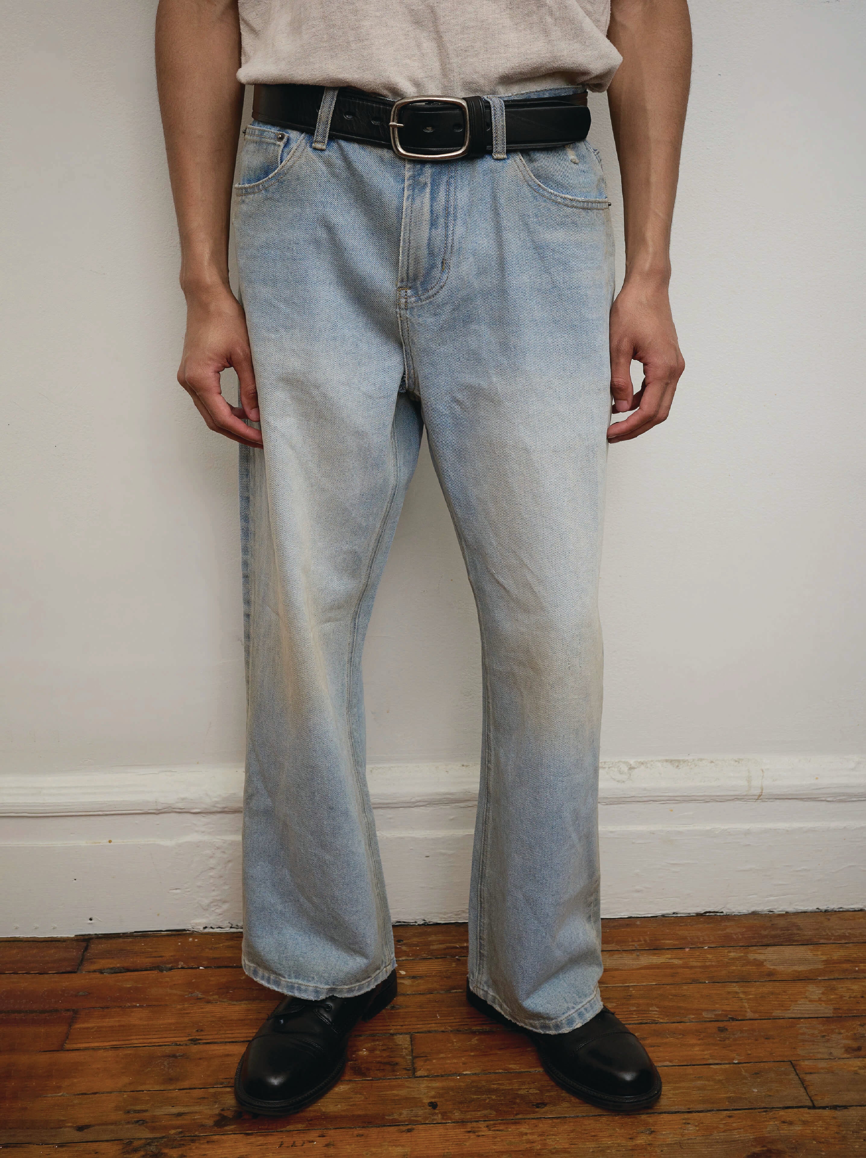 A person stands indoors wearing Found Flare Jeans in light blue denim with a black belt, a tucked-in light shirt, and black shoes; only the lower half of the body is shown.