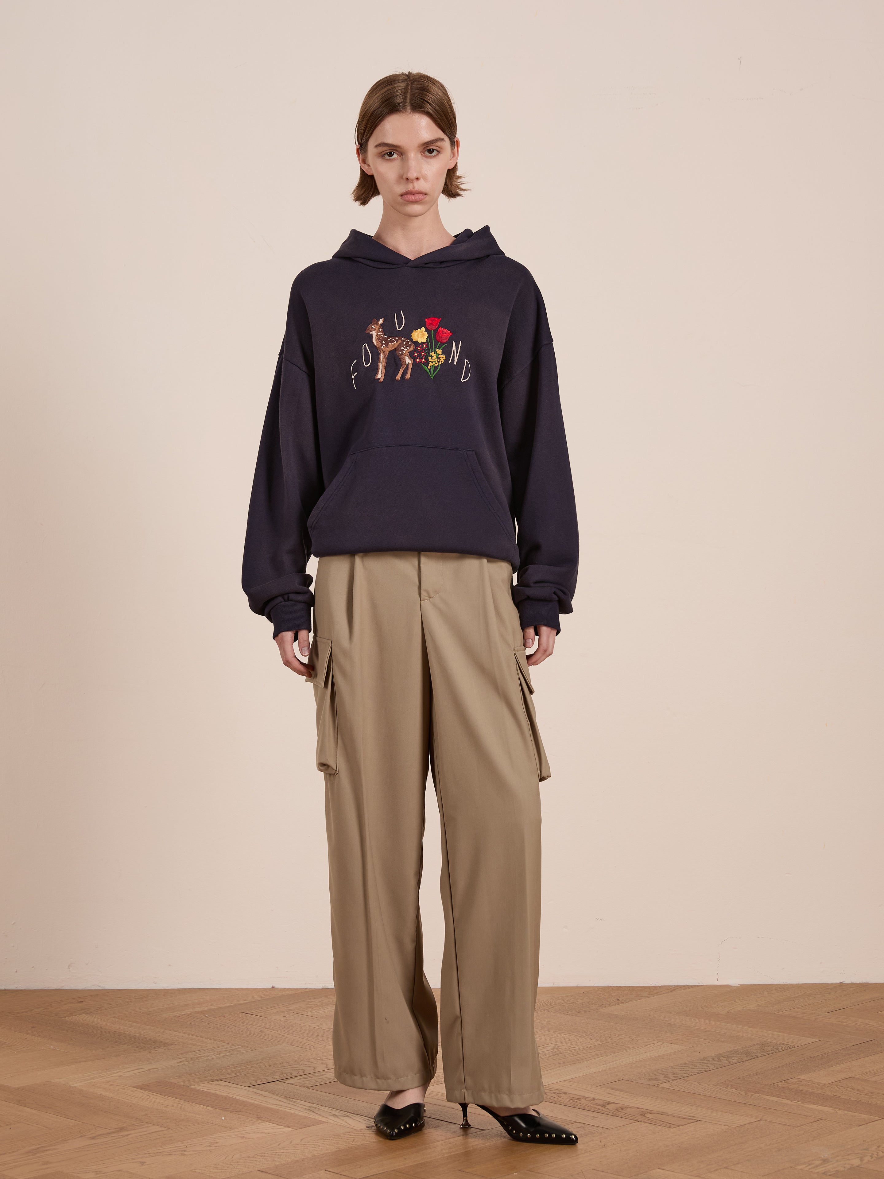 Indoors, someone wears a FOUND Floral Embroidered Deer Hoodie in navy, made from vintage-washed French terry cotton for a unisex relaxed fit. It’s paired with beige wide-legged pants and black shoes on a wooden floor.