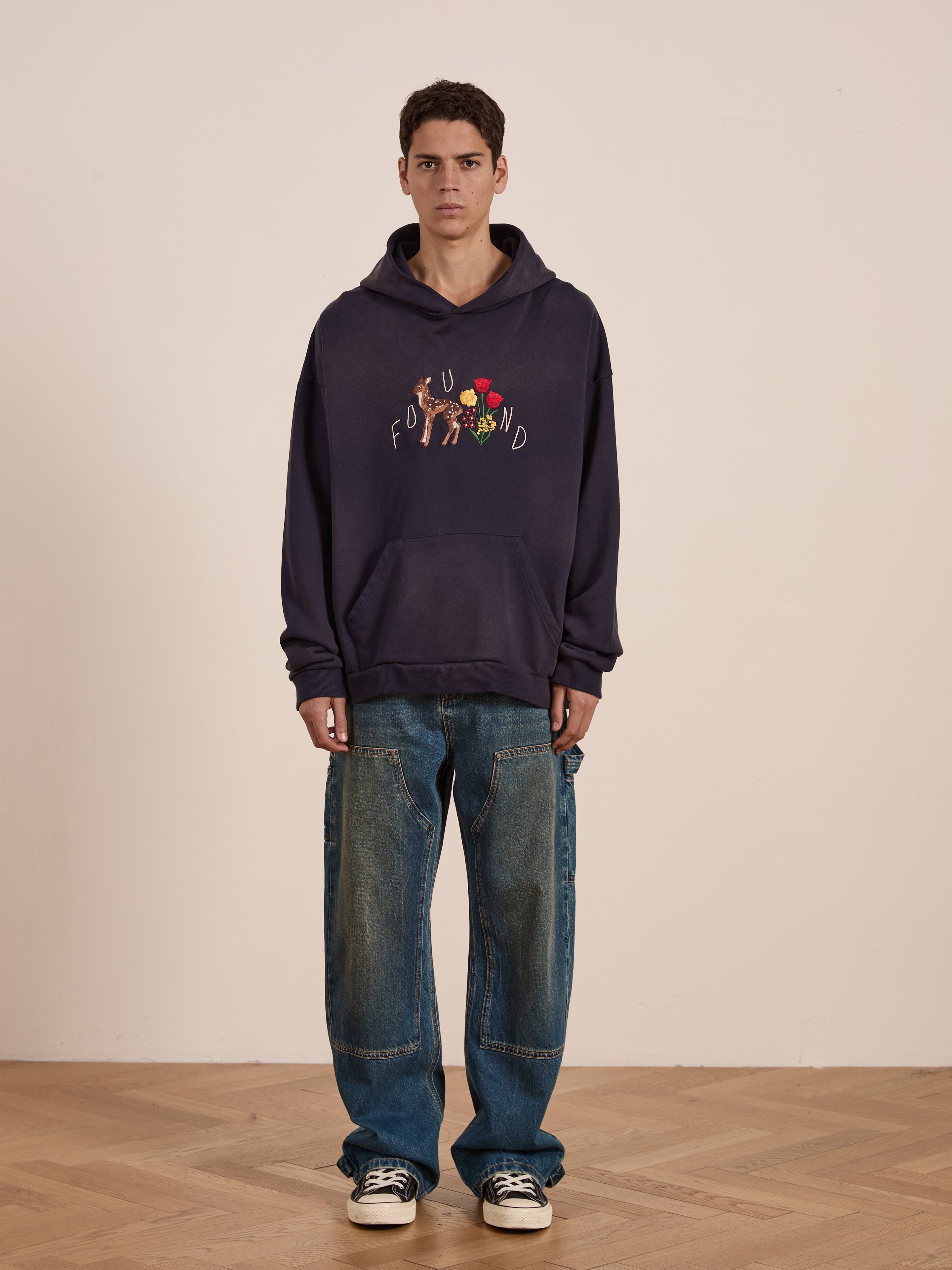 A person in FOUND's Floral Embroidered Deer Hoodie made from vintage-washed French terry cotton, paired with loose jeans and black sneakers, stands on a wooden floor against a plain background. The unisex relaxed fit offers effortless style.