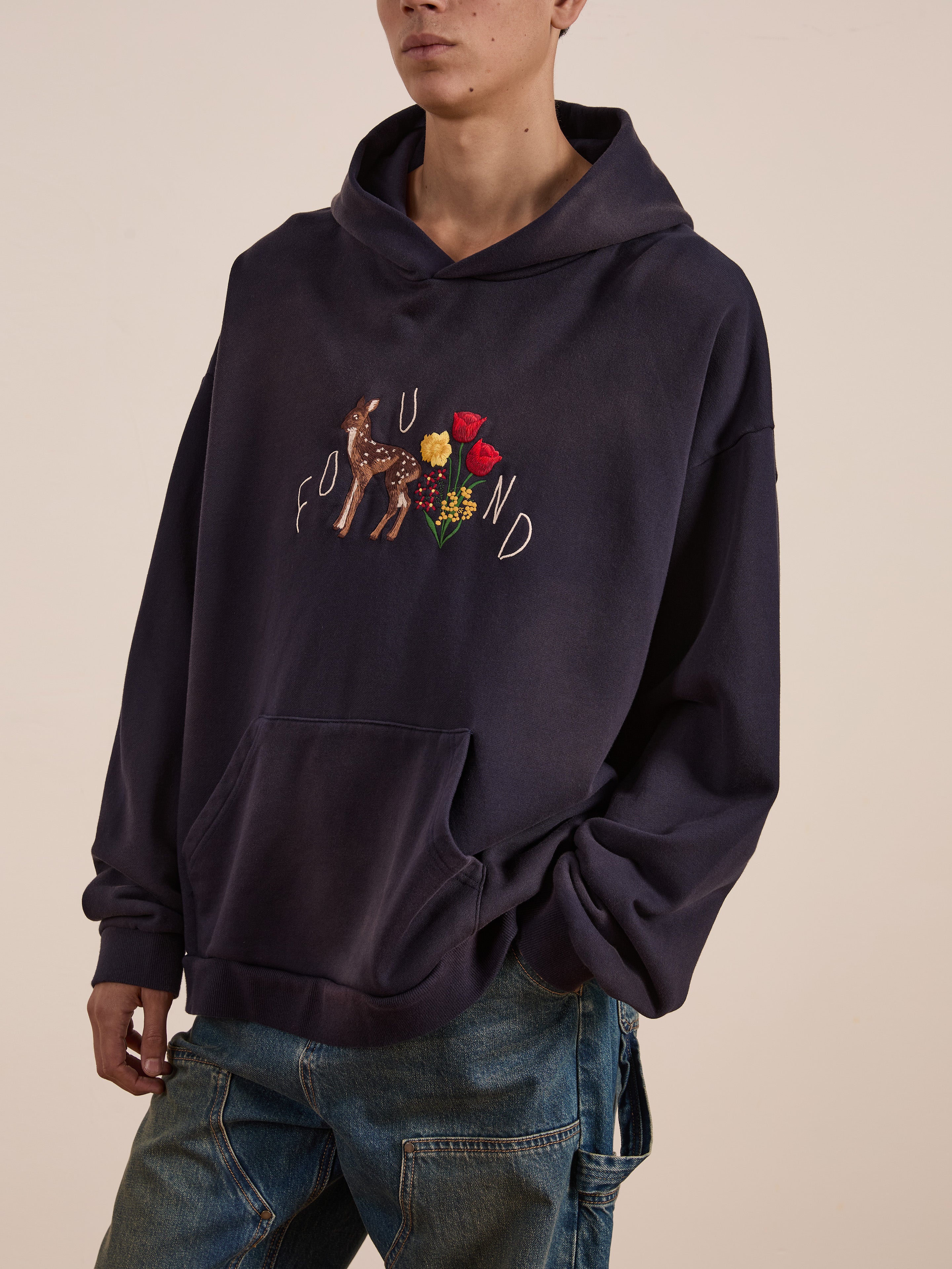 Wearing the unisex Floral Embroidered Deer Hoodie by FOUND, featuring vintage-washed French terry cotton with floral and deer motifs, pairs perfectly with blue jeans.