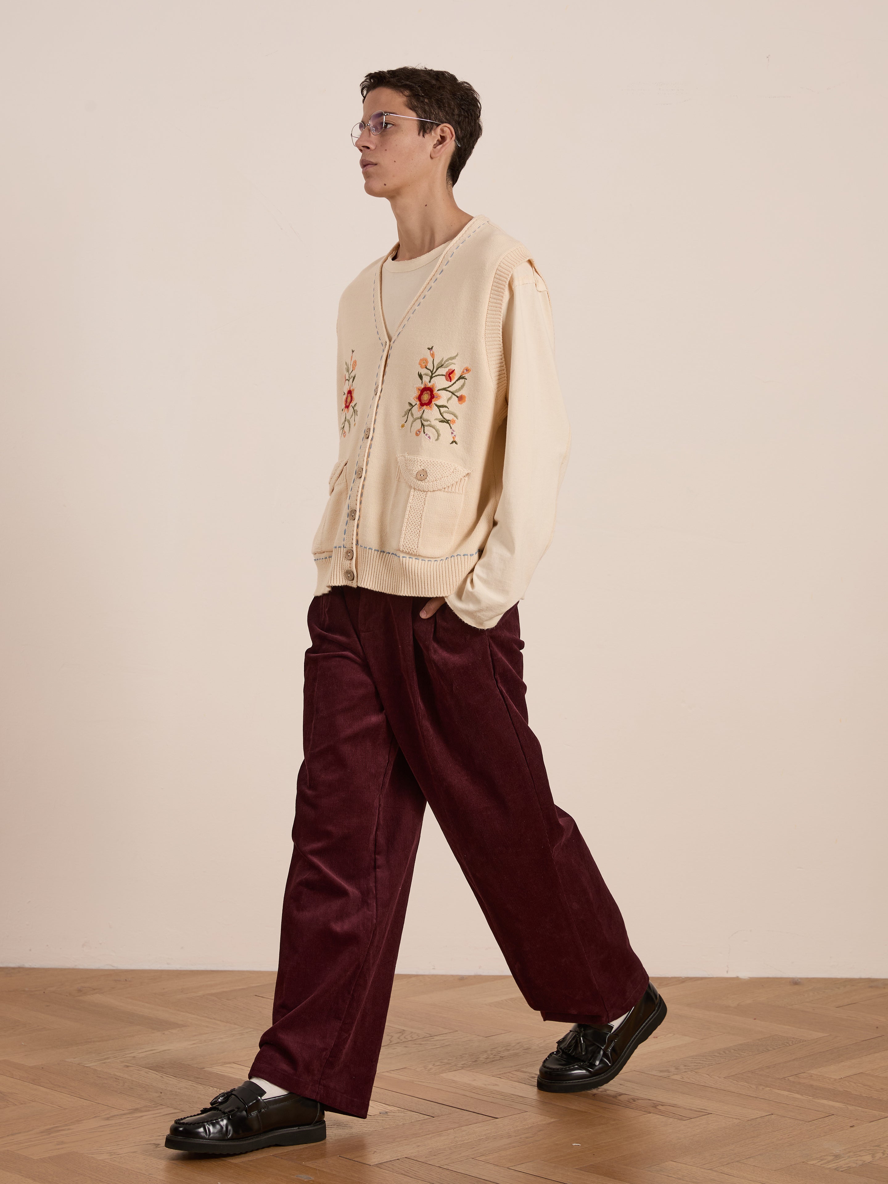 Dressed in a retro-style Floral Embroidered Knit Vest by FOUND over a white shirt, with burgundy wide-leg pants and black shoes, a person walks elegantly on a wooden floor.