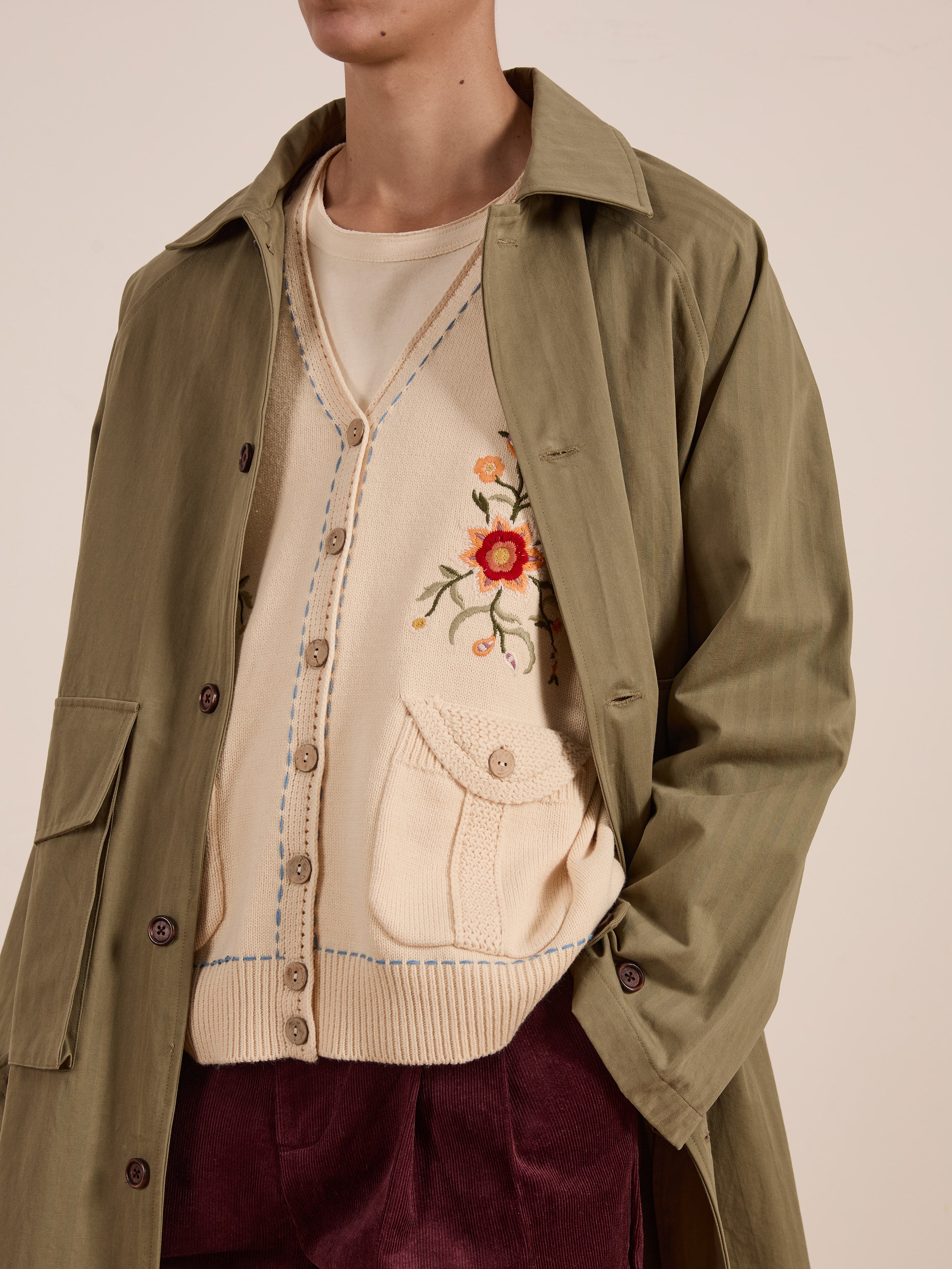 A person wears a vintage-style outfit with a green coat over a white shirt and the "Floral Embroidered Knit Vest" by FOUND. They finish the look with maroon pants, adding unisex appeal to their style.