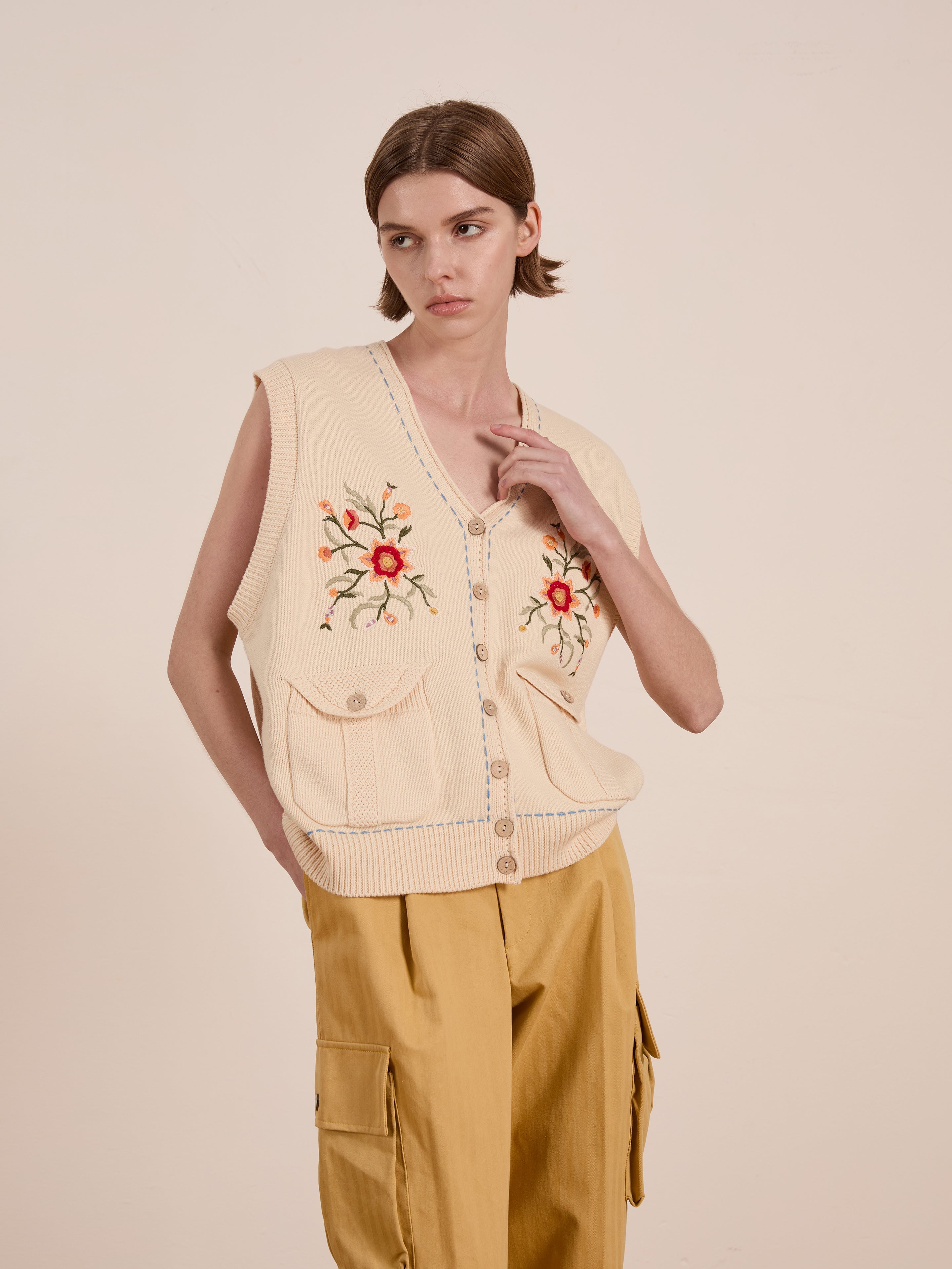 Against a plain backdrop, an individual showcases a vintage-inspired aesthetic in FOUND's unisex beige Floral Embroidered Knit Vest, paired stylishly with mustard cargo pants.