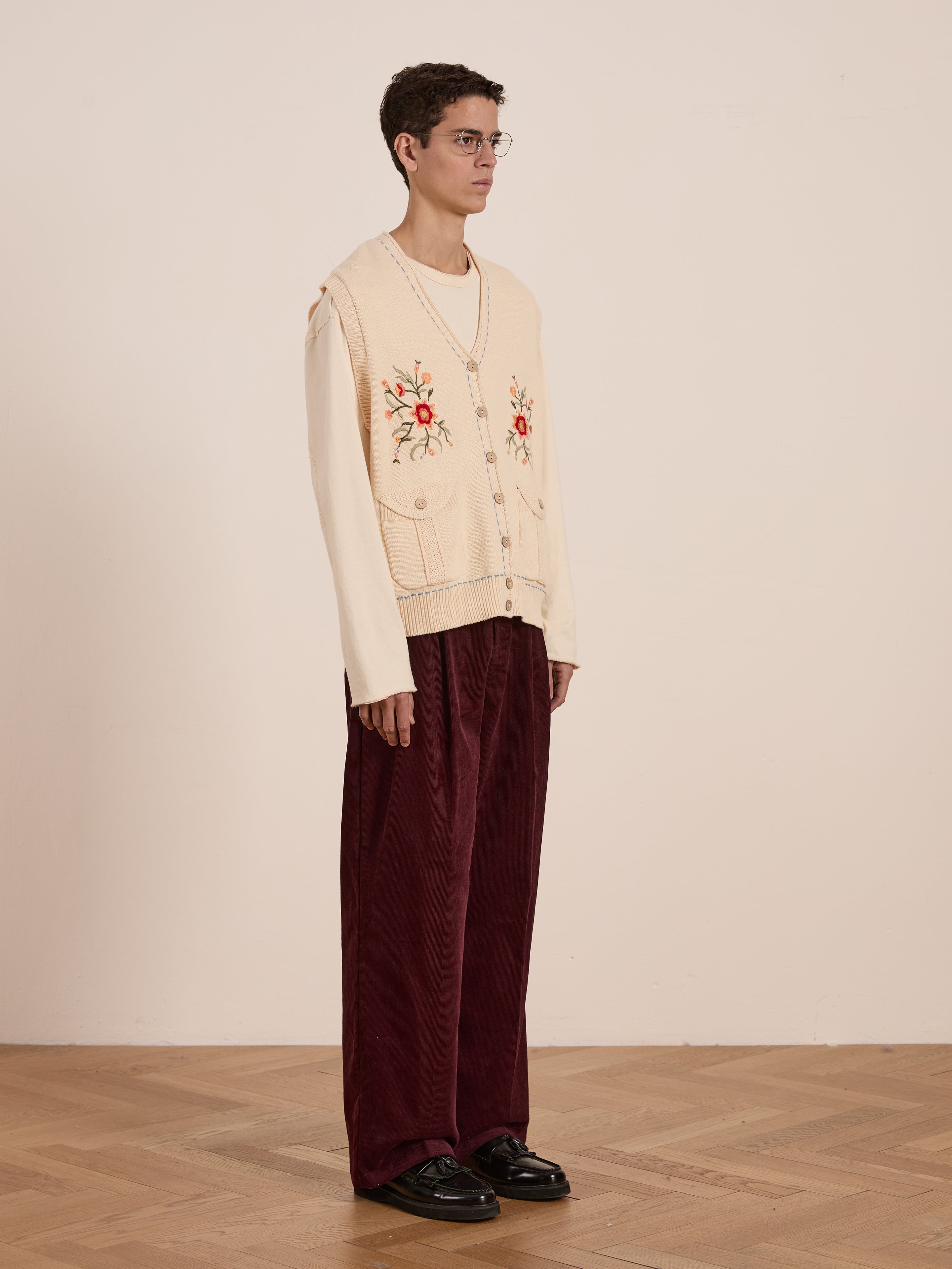 A person wears glasses, standing on a wooden floor against a plain wall, styled in a vintage-inspired Floral Embroidered Knit Vest by FOUND over a long-sleeve shirt and wide maroon pants, exuding an effortlessly unisex look.