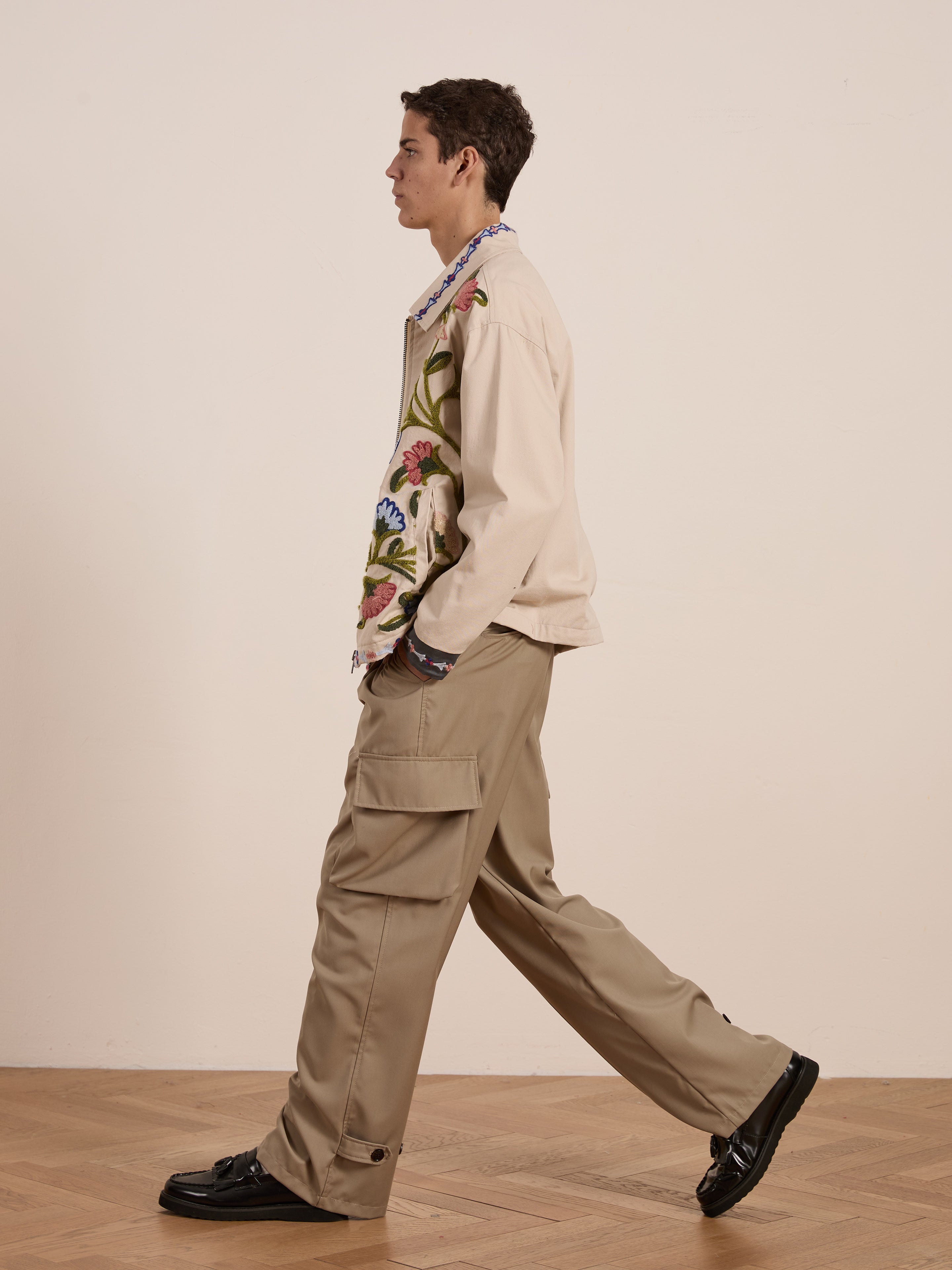 A person in profile walks indoors, wearing FOUND's unisex Floral Embroidered Work Jacket with a relaxed fit, paired with khaki cargo pants. Black shoes tread softly on the wooden floor against a plain backdrop.