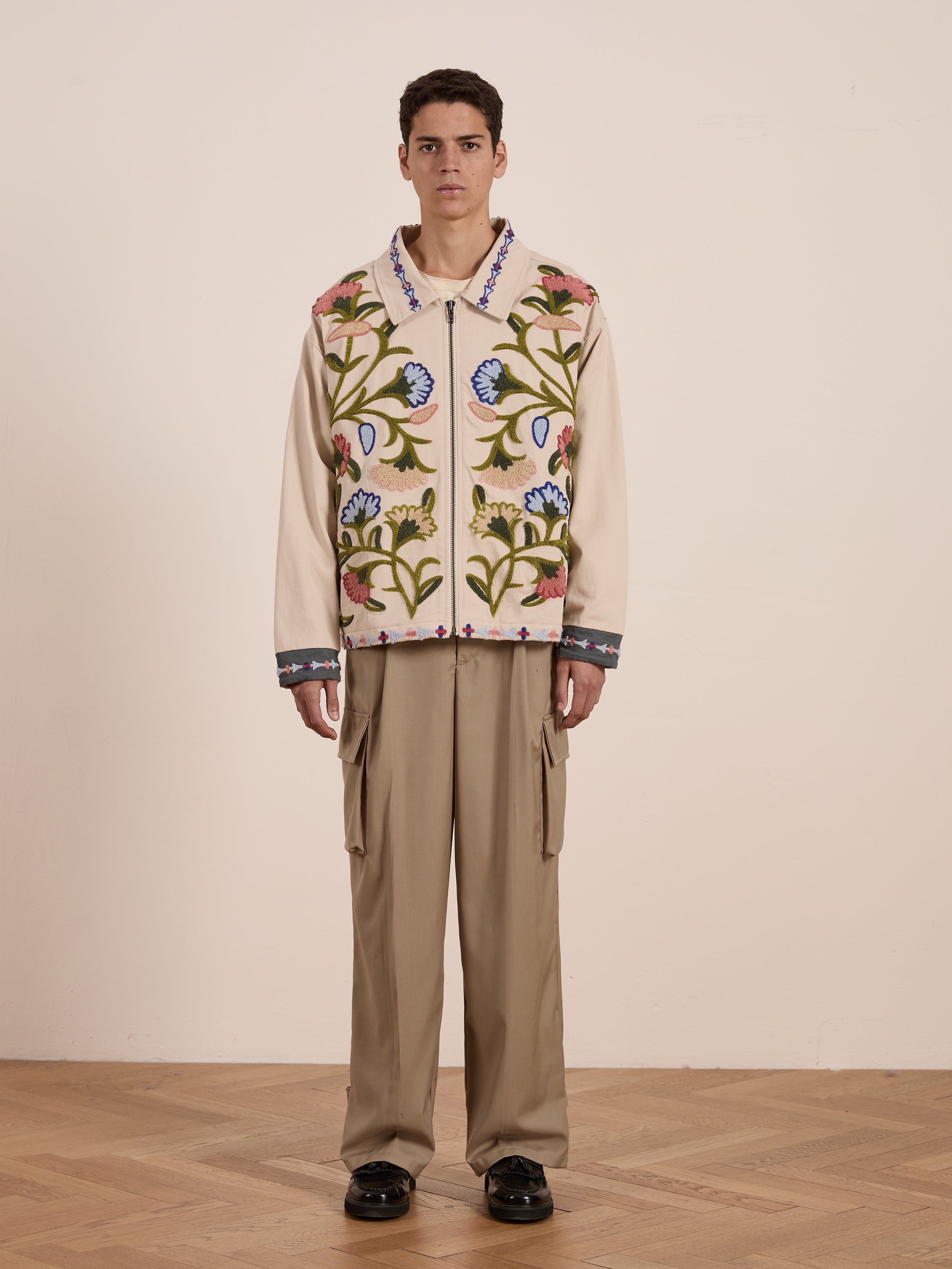 A person in a unisex relaxed fit, FOUND's Floral Embroidered Work Jacket paired with khaki cargo pants stands on a wooden floor against a plain background.