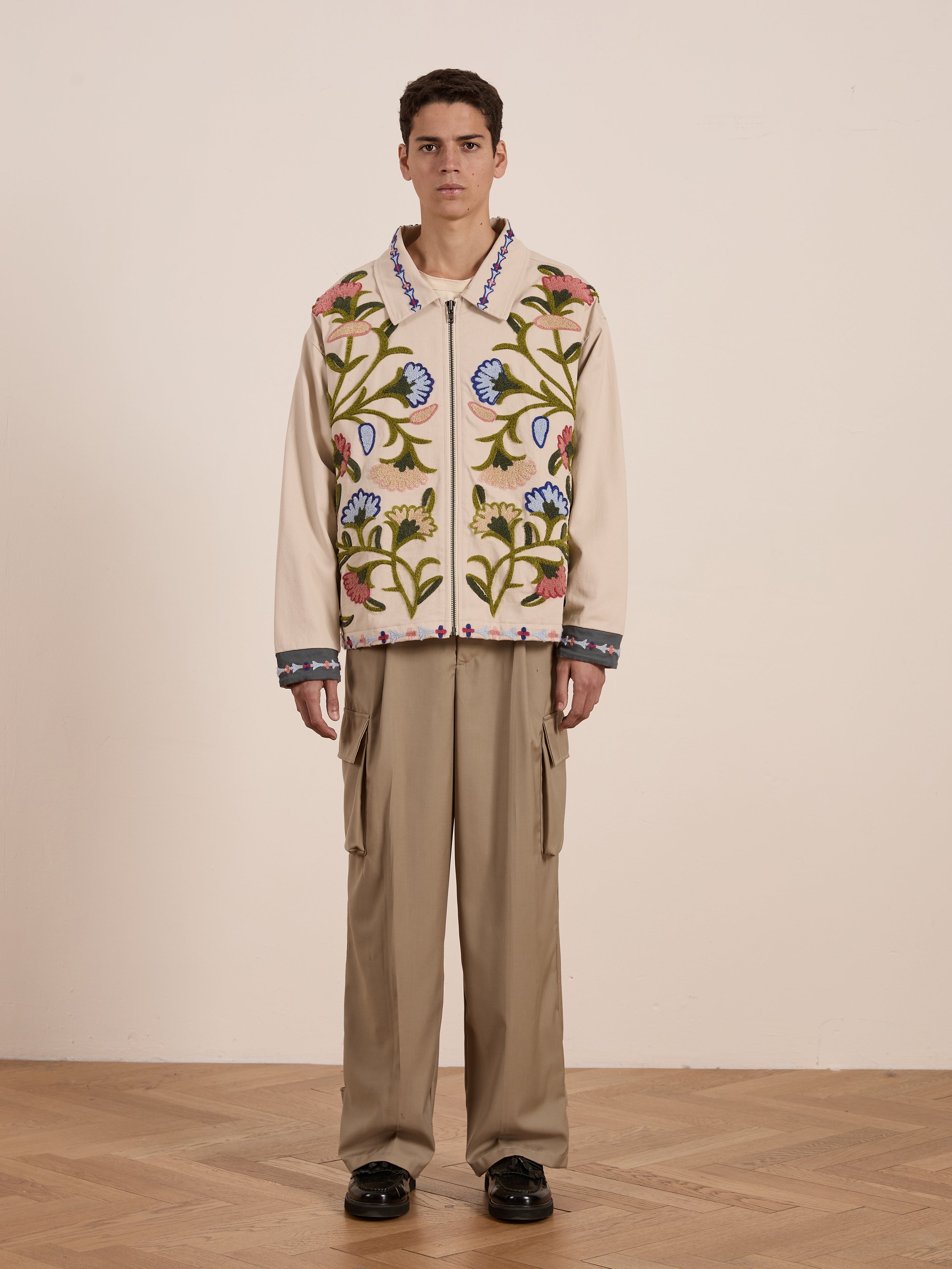 A person stands on a wooden floor against a plain wall, wearing the FOUND Floral Embroidered Work Jacket, a unisex beige cotton canvas piece with colorful floral embroidery, paired with tan cargo pants for an effortlessly stylish and comfortable look.