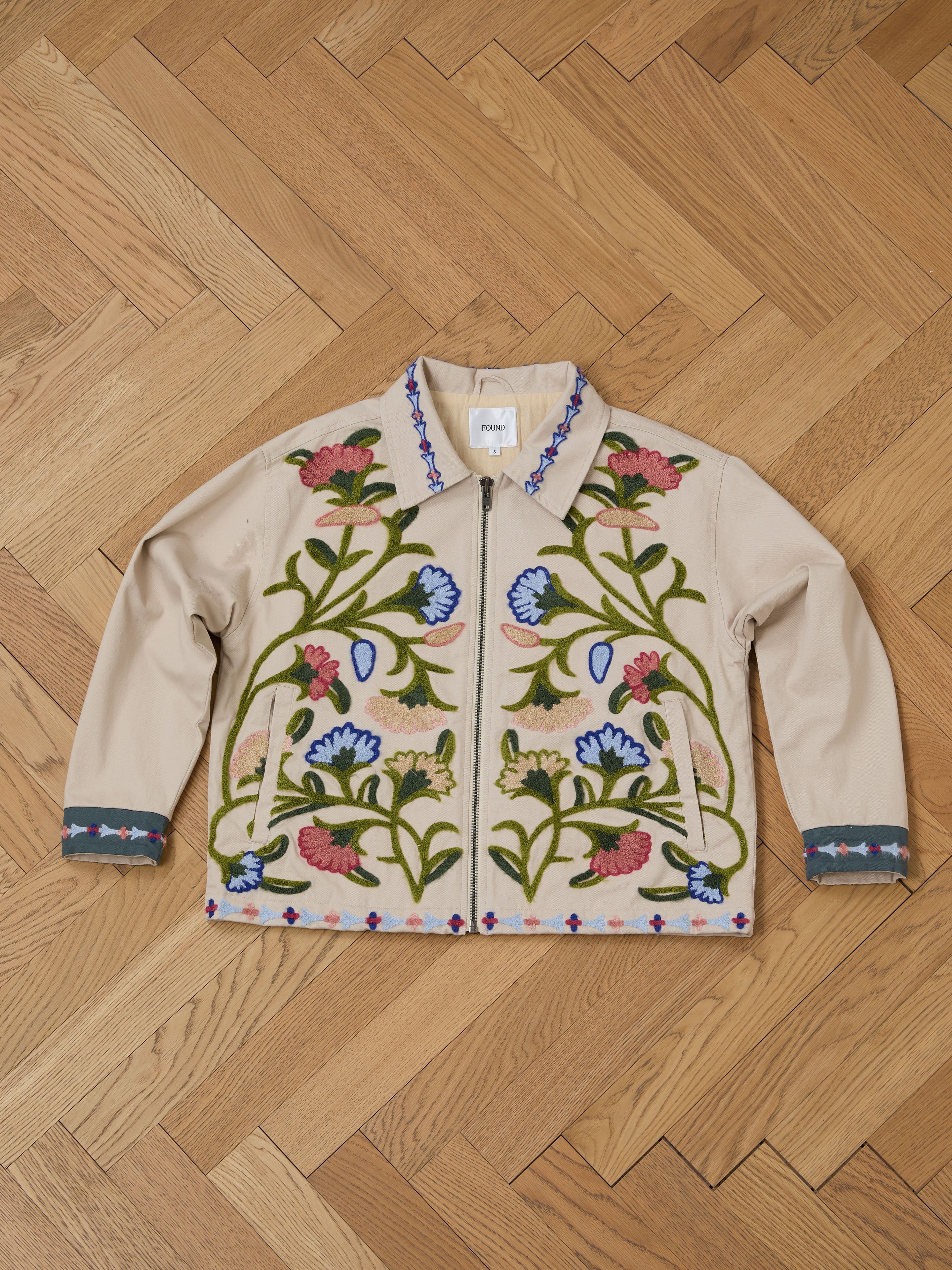 FOUND's Floral Embroidered Work Jacket, a unisex relaxed-fit piece made from soft beige cotton canvas, rests on a herringbone wood floor.