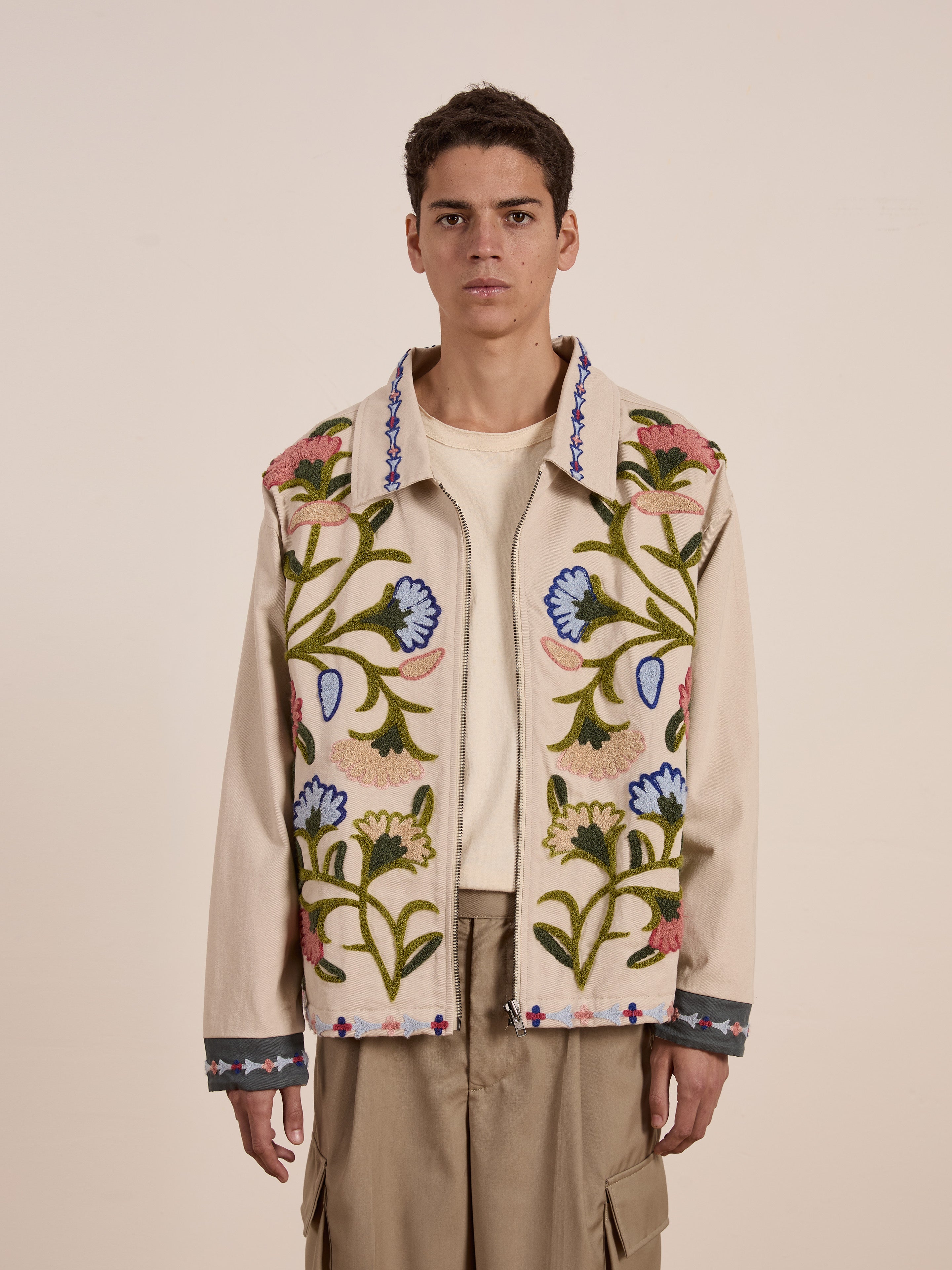 A person wears FOUND's unisex Floral Embroidered Work Jacket in beige cotton canvas, with vibrant floral embroidery that contrasts nicely against a plain shirt and khaki pants against a neutral backdrop.