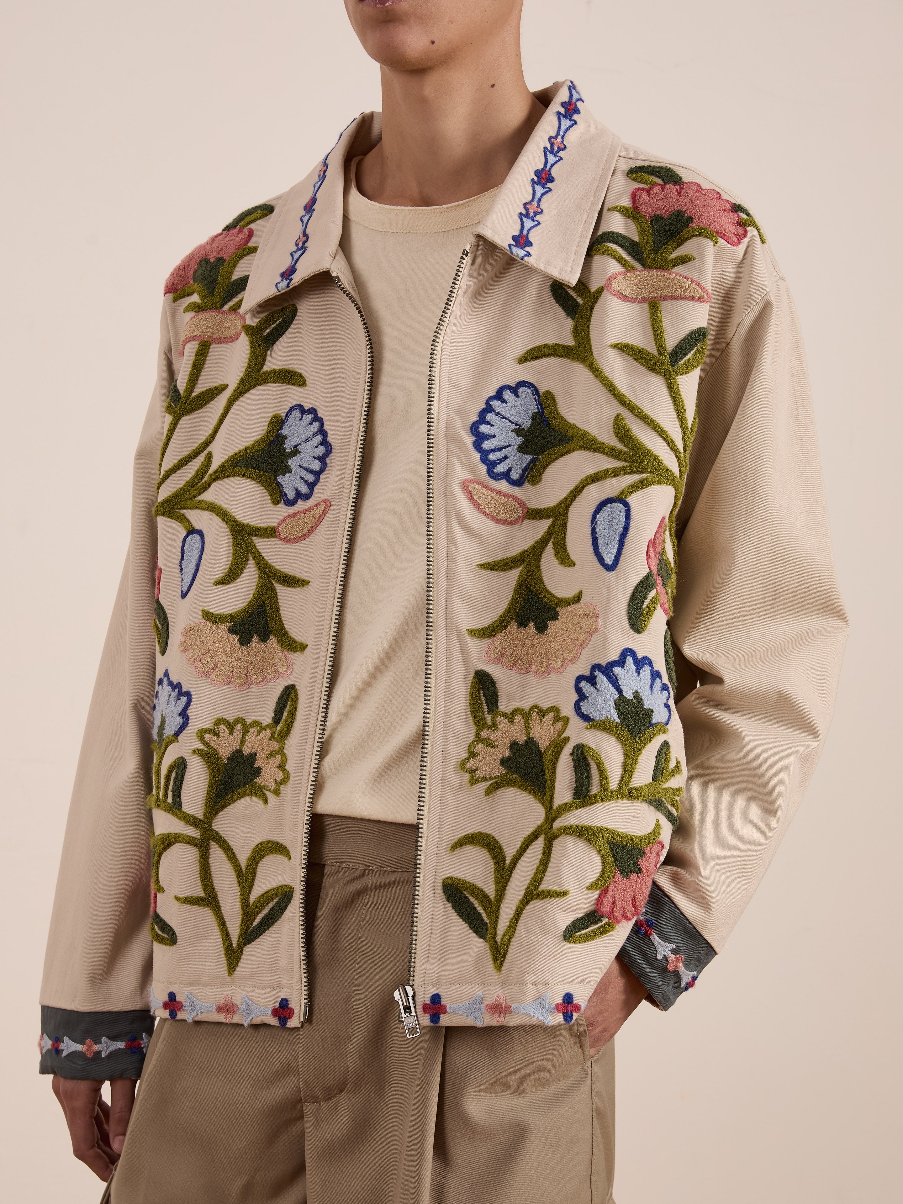 A person is wearing FOUND's beige cotton canvas Floral Embroidered Work Jacket in a unisex relaxed fit over a light shirt and tan pants.