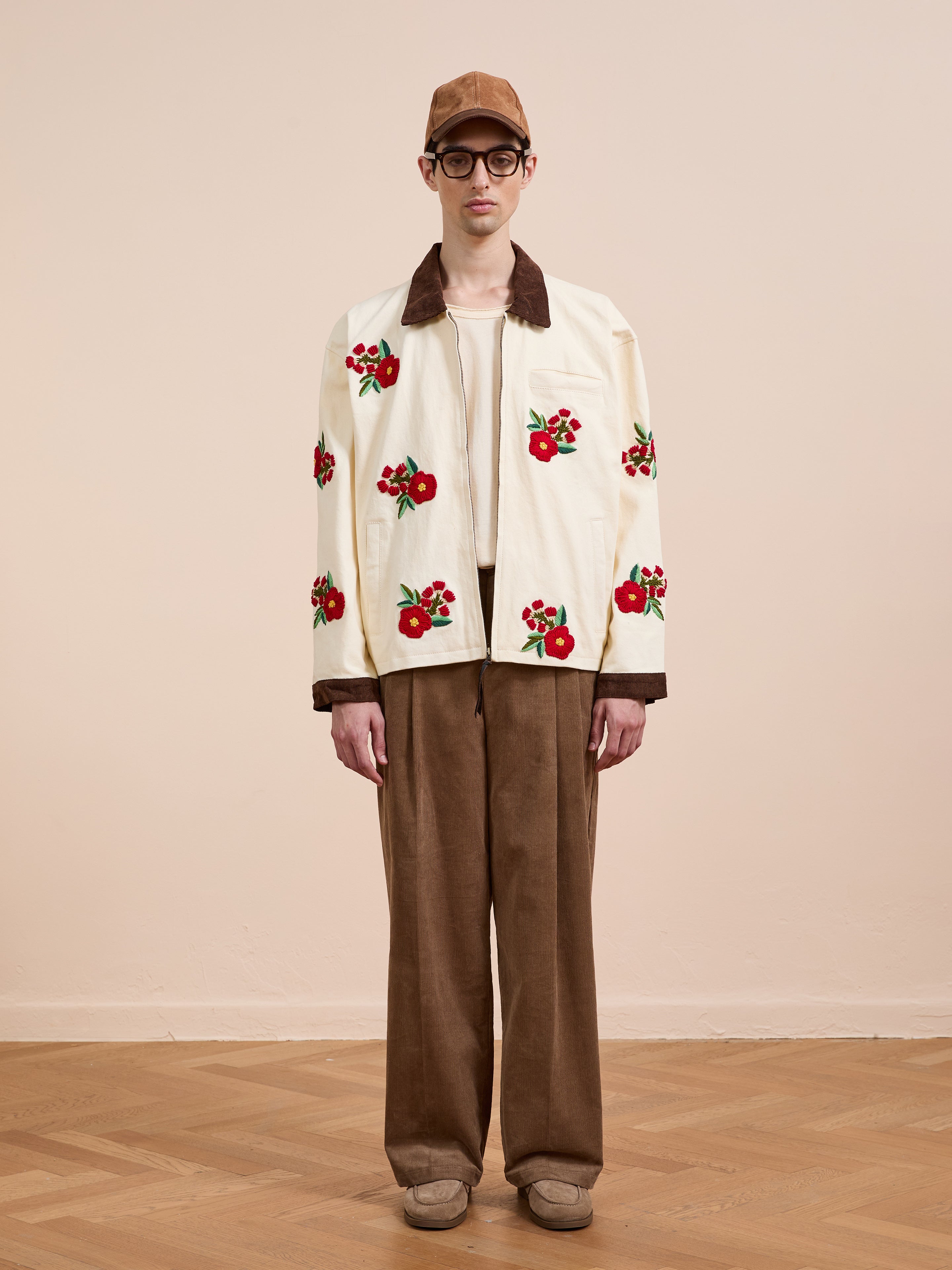A person stands indoors against a beige background wearing the Found Floral Hand Embroidered Work Jacket, wide brown corduroy pants, a brown cap, glasses, and beige shoes.