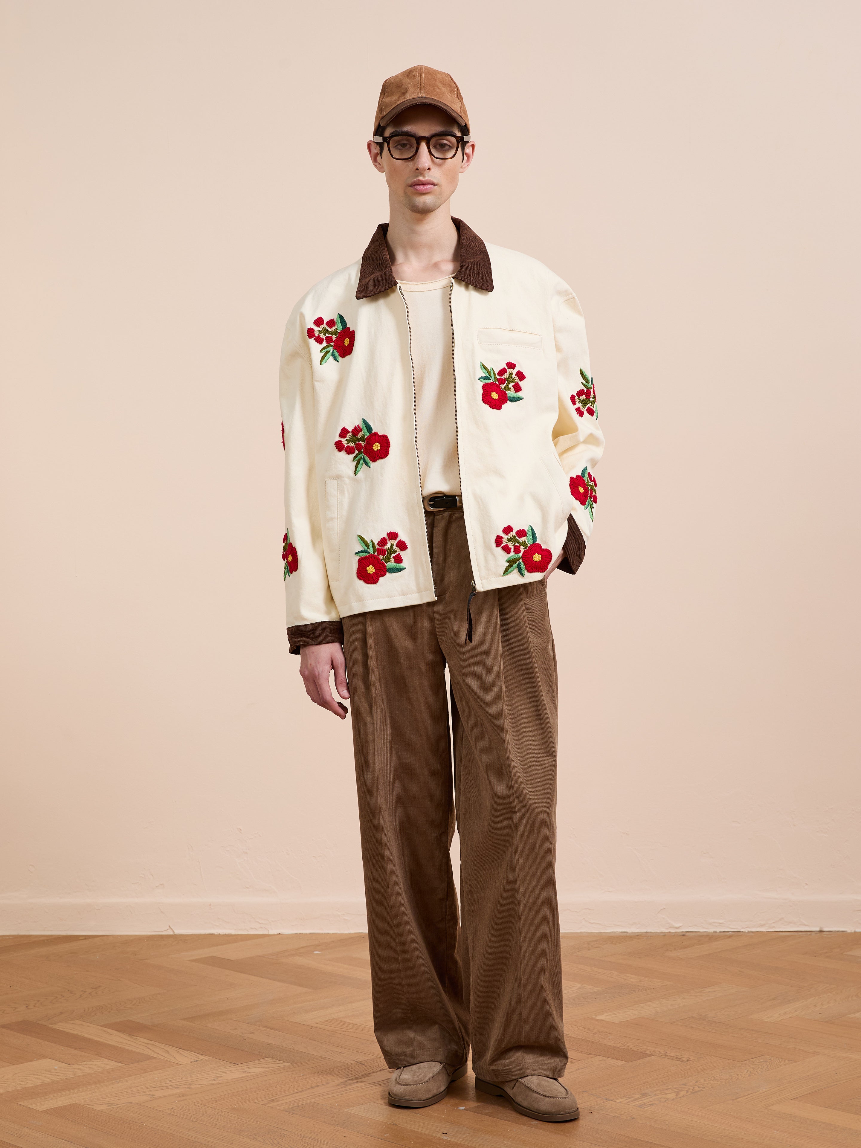 A person stands in a neutral room wearing a beige cap, glasses, the Found Floral Hand Embroidered Work Jacket, a white shirt, wide brown pants, and beige shoes.