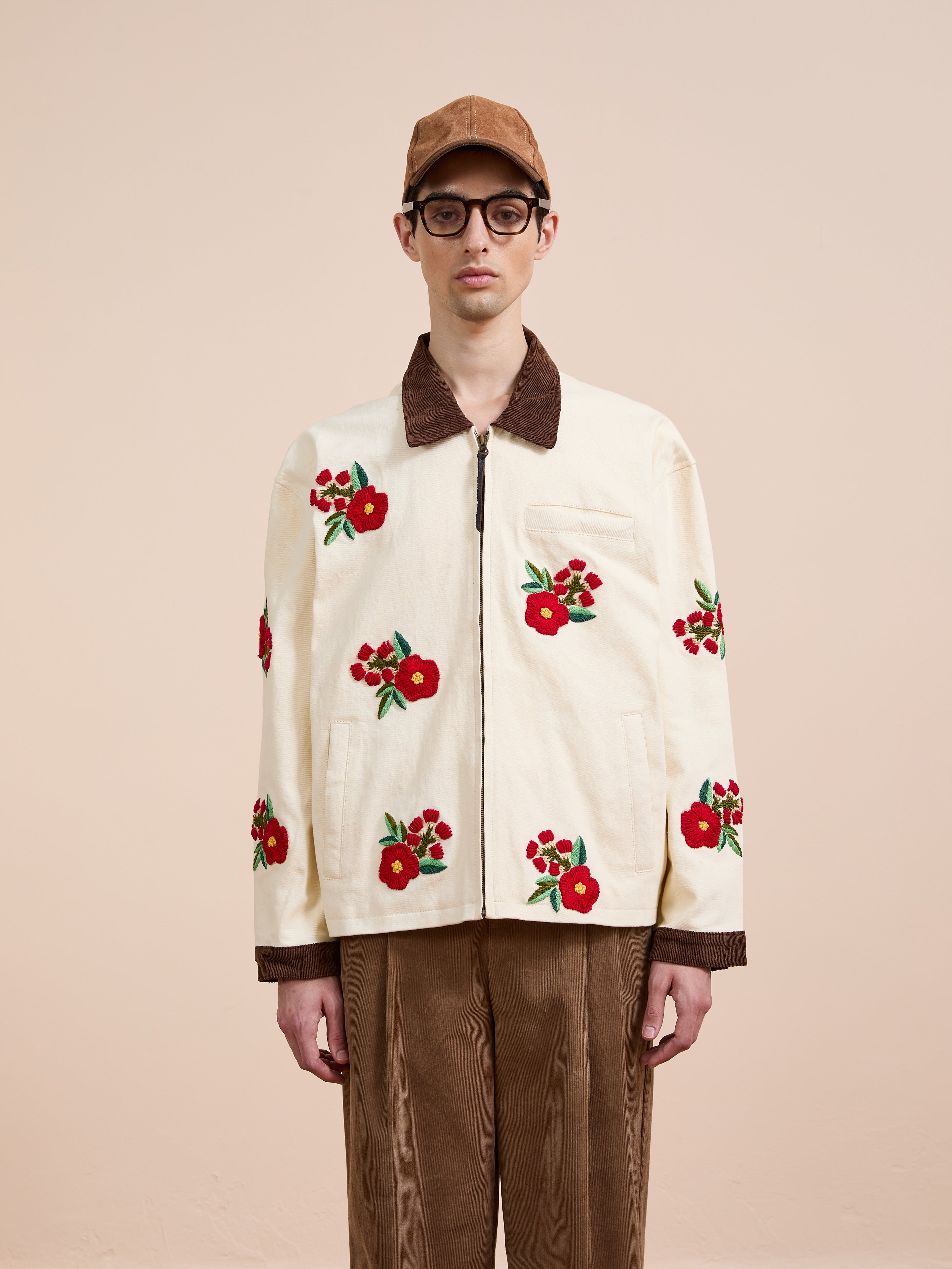 A person in a tan cap and glasses models the Found Floral Hand Embroidered Work Jacket, inspired by vintage South Asian designs, against a plain beige background.