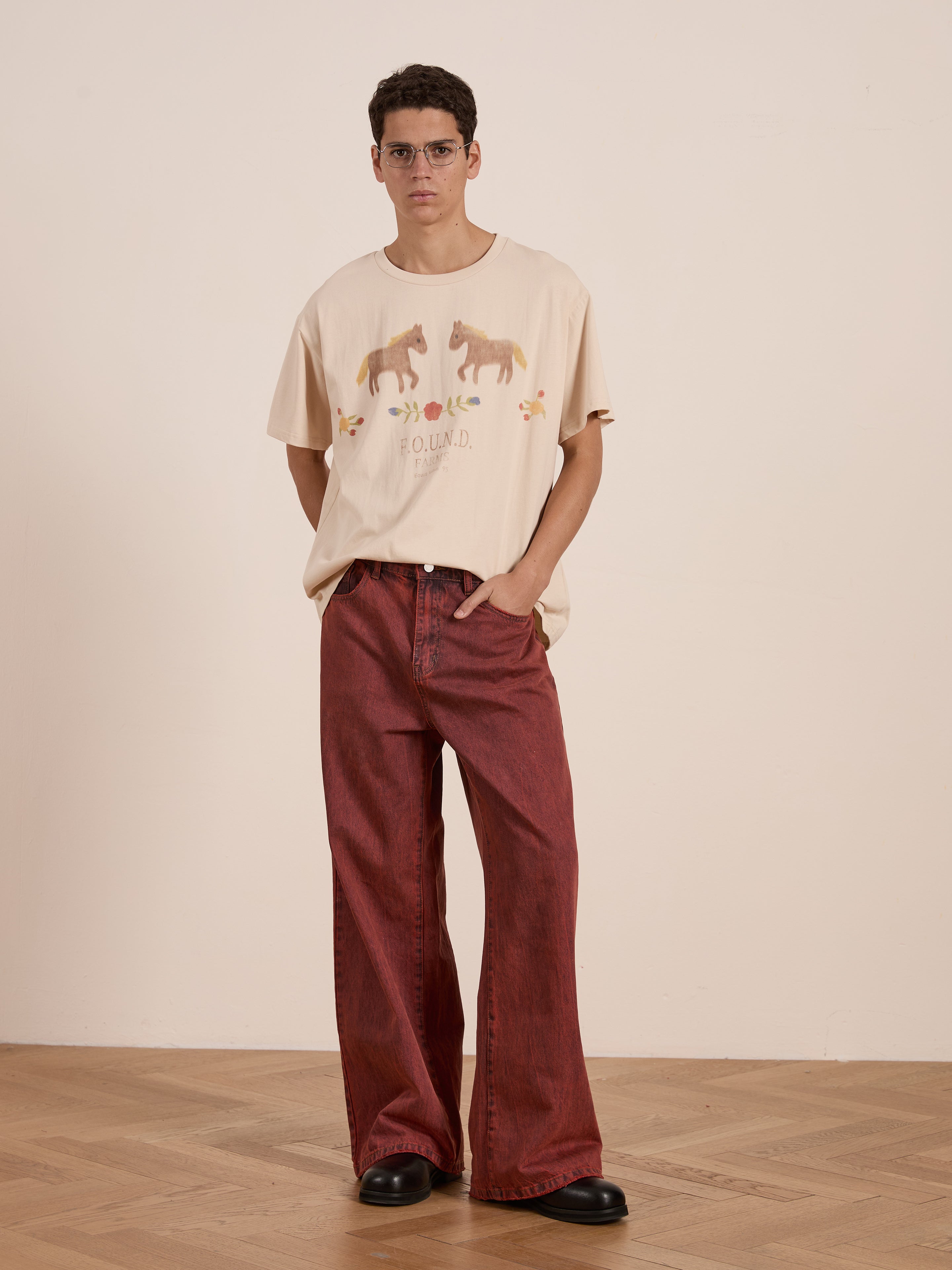 A person with glasses wearing a FOUND Floral Horse Farm Tee featuring equestrian graphics, wide red pants, and black shoes stands on a wooden floor against a plain beige wall.