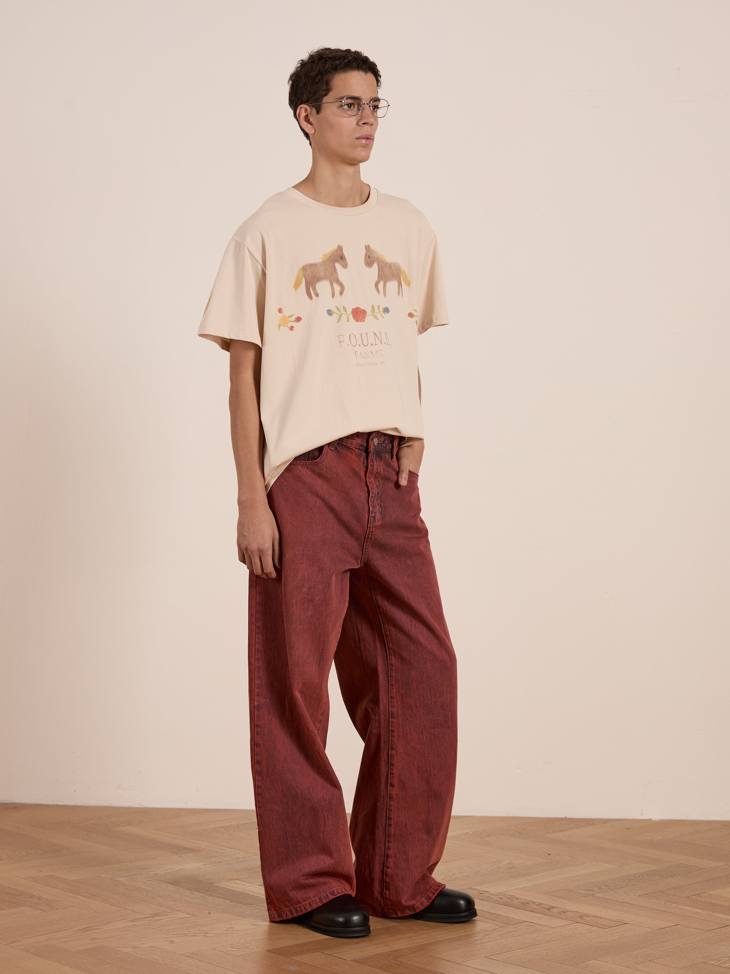 A person in glasses wears FOUND's Floral Horse Farm Tee featuring equestrian graphics, paired with red wide-leg pants, standing on a wooden floor against a plain wall.