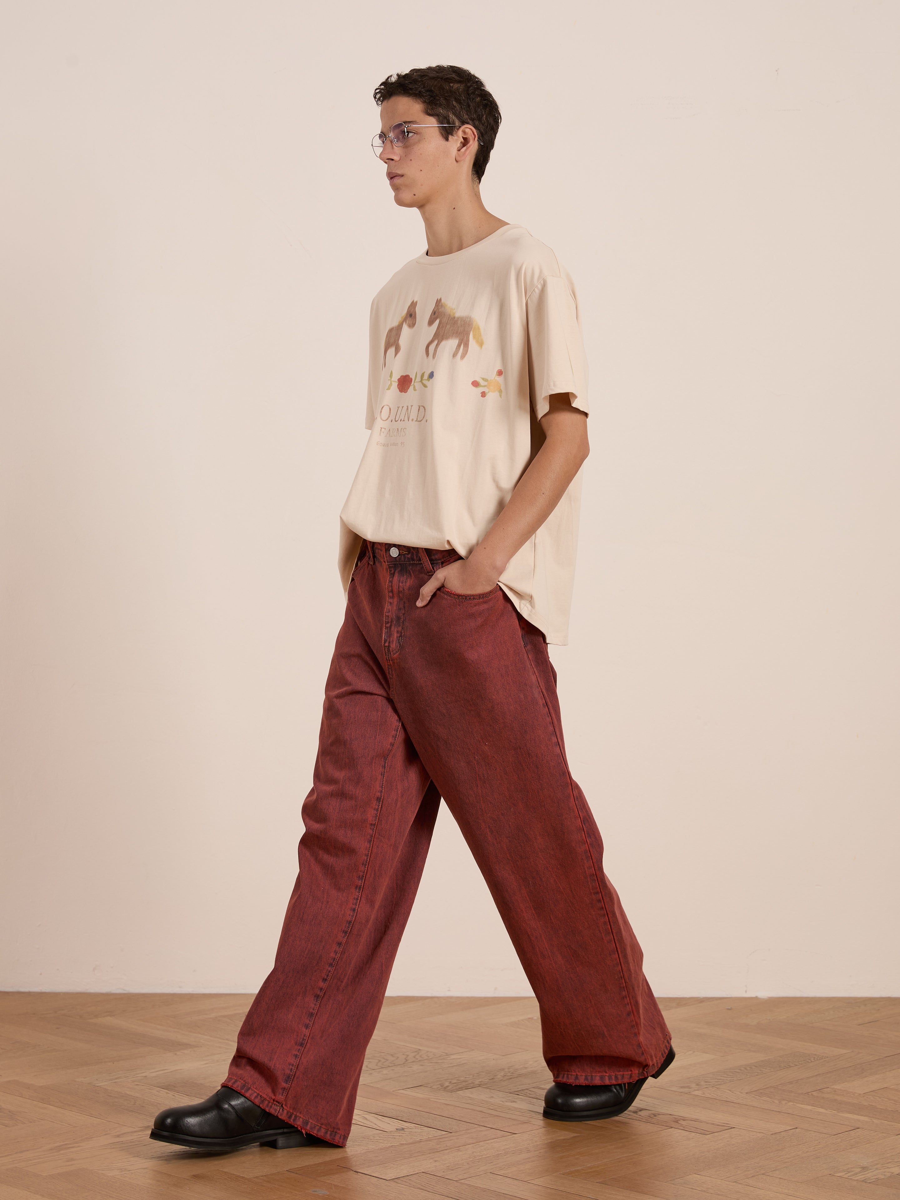 Wearing FOUND's Floral Horse Farm Tee with equestrian graphics, beige in color, and matched with red wide-leg pants, black shoes, and glasses. They pose elegantly on a wooden floor against a plain wall, radiating vintage artistry.