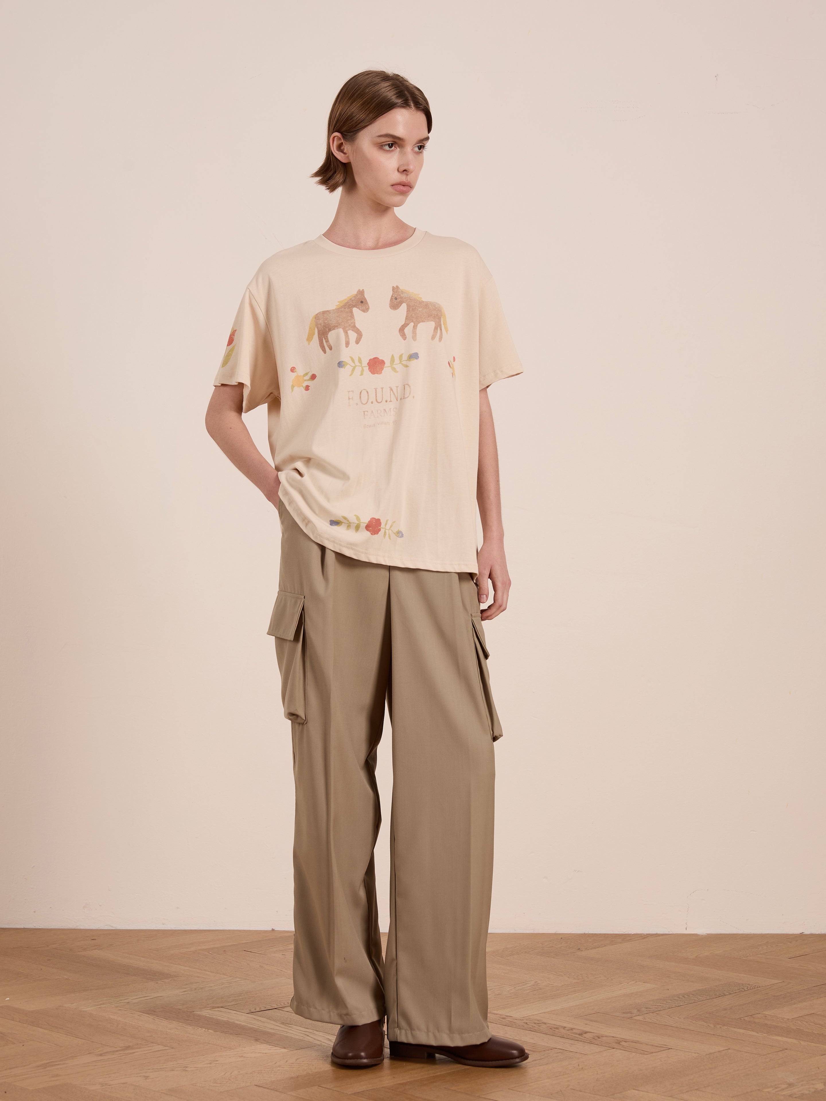 A person stands on a wooden floor, wearing FOUND's Floral Horse Farm Tee in beige and light brown cargo pants.