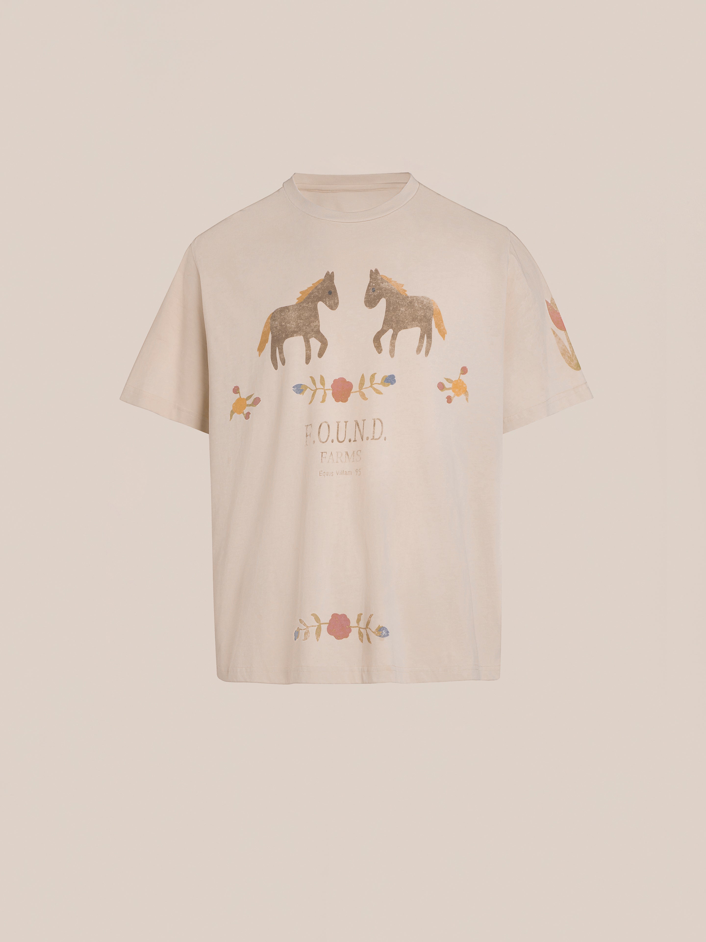 The Floral Horse Farm Tee by FOUND is a beige T-shirt showcasing countryside-inspired equestrian graphics. It features two horse illustrations with floral accents on the chest and "ROUND FARM" text underneath.