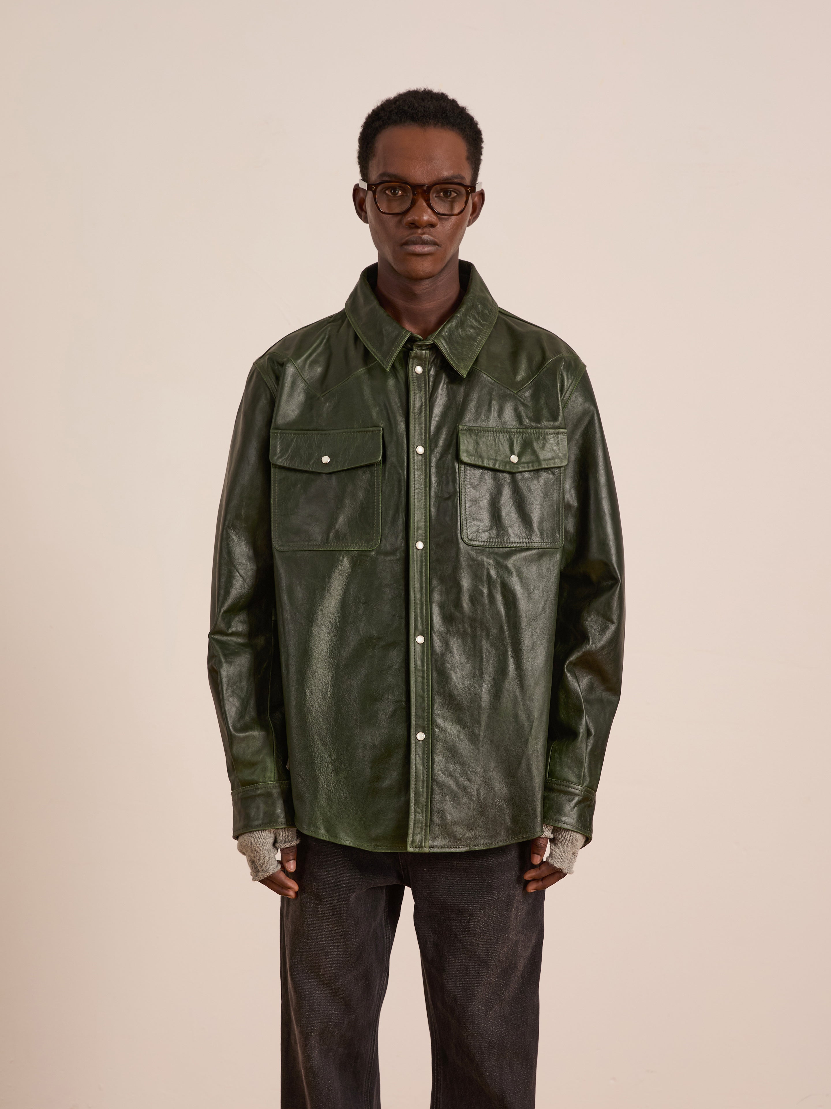 A person wearing glasses, dark pants, fingerless gloves, and the FOUND Forest Leather Western Shirt, a dark green unisex style with chest pockets, stands against a plain light background.