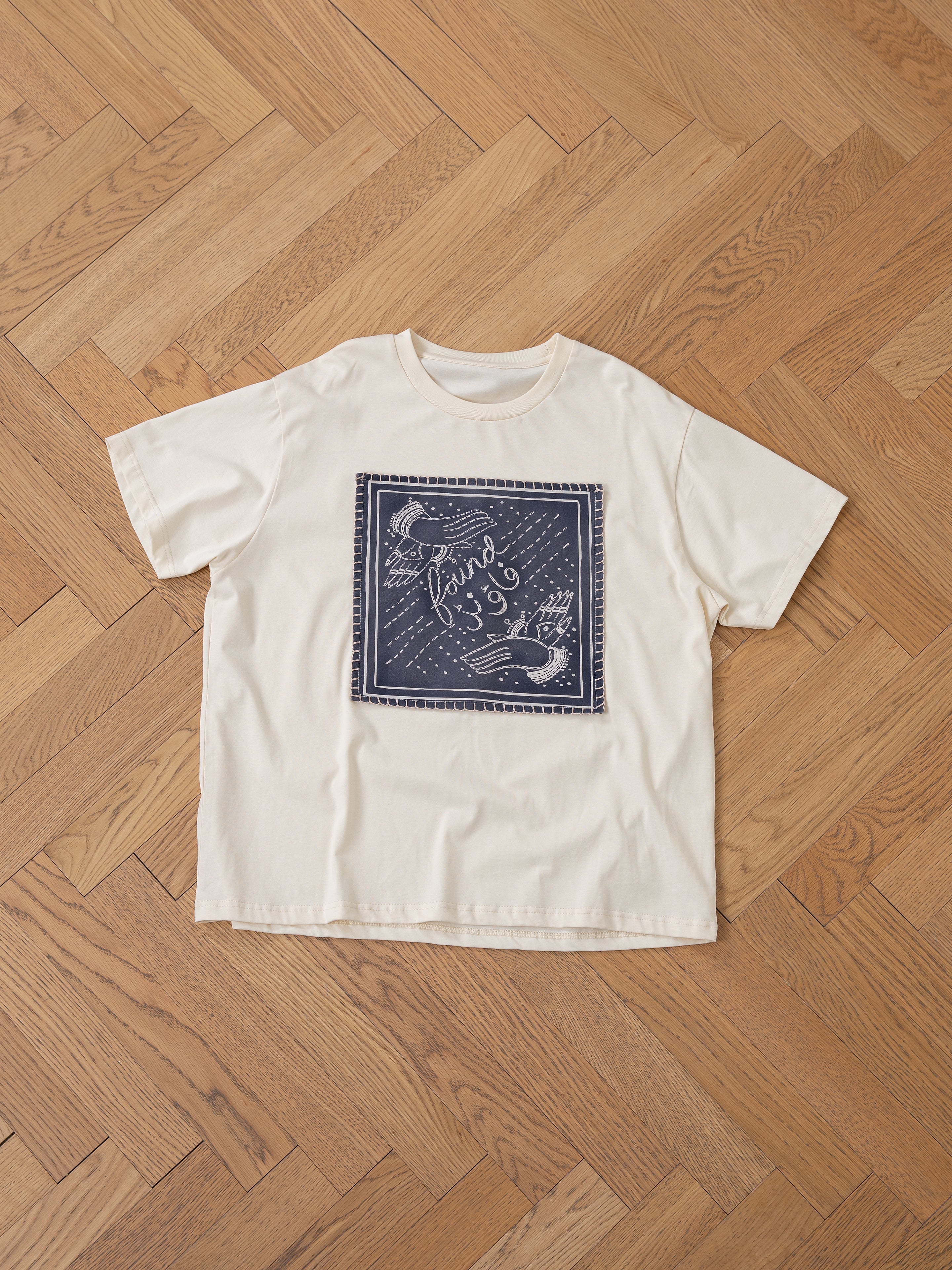 The Urdu Graphic Tee by FOUND is a beige T-shirt featuring a navy blue square graphic of birds and Urdu cursive text, inspired by Mughal paintings, displayed flat on a wooden floor.