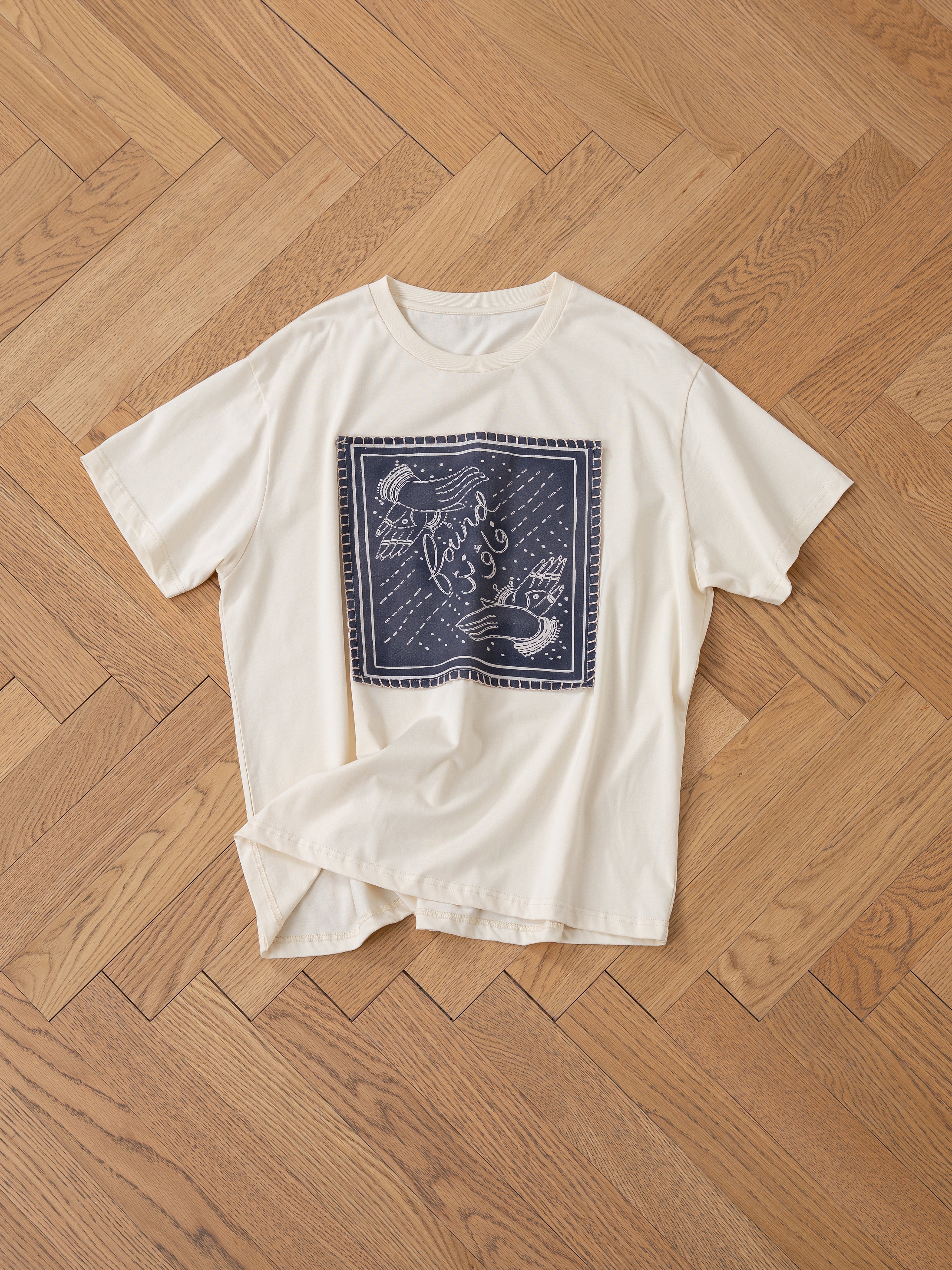 The FOUND Urdu Graphic Tee, a beige T-shirt featuring a square navy graphic patch adorned with fish and the words "friend from sea," echoing the style of South Asian Mughal paintings, is elegantly displayed on a wooden parquet floor.