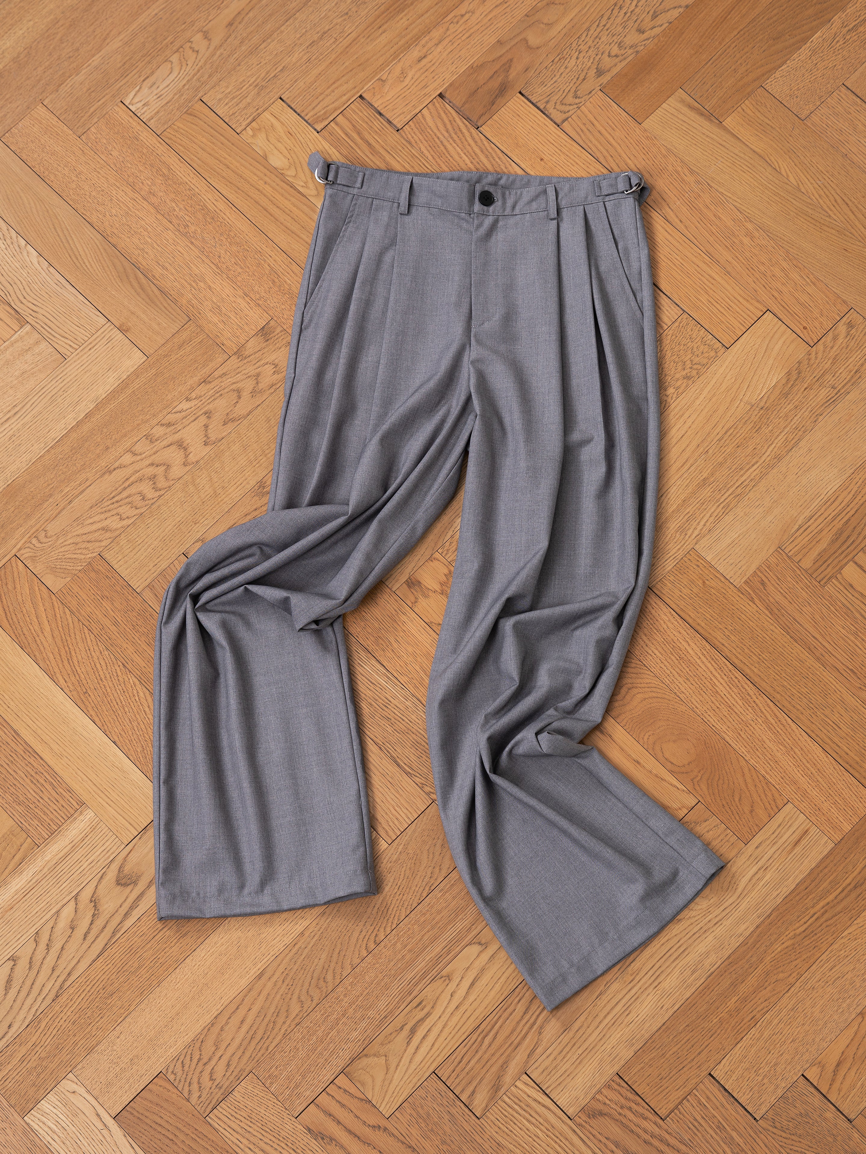 FOUND's Pleated Trousers, featuring a sophisticated wide-leg high-rise design in gray, are gracefully displayed on a wooden herringbone floor.