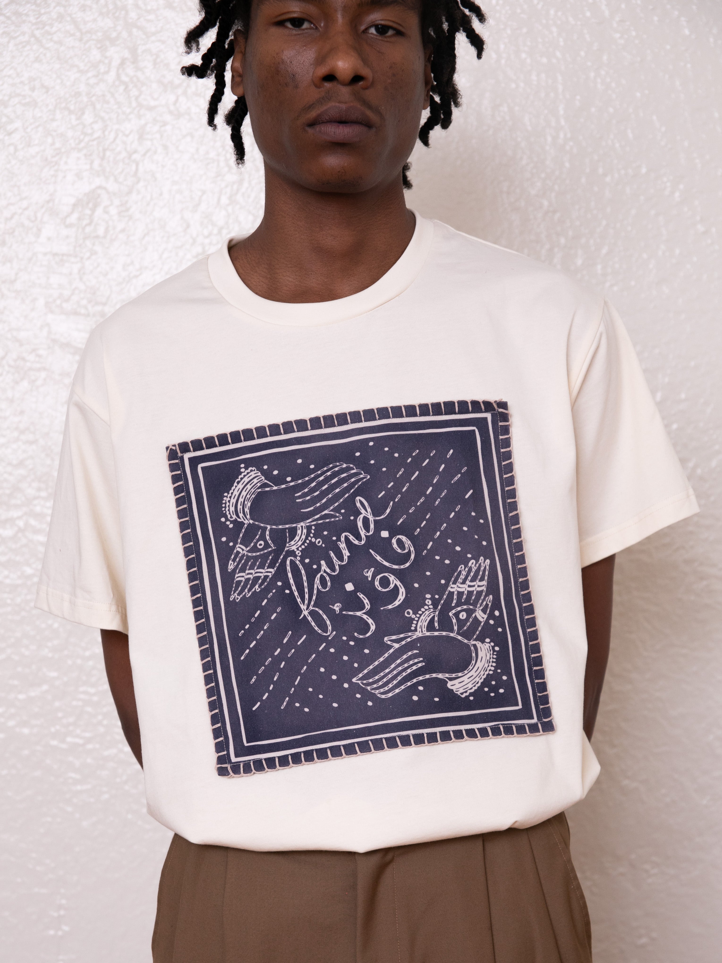 A person wearing the Hand Art Graphic Tee by FOUND, featuring an Urdu graphic in navy blue with tilted hands and cursive text on a white t-shirt, is paired with cotton twill brown pants and stands against a light textured wall.