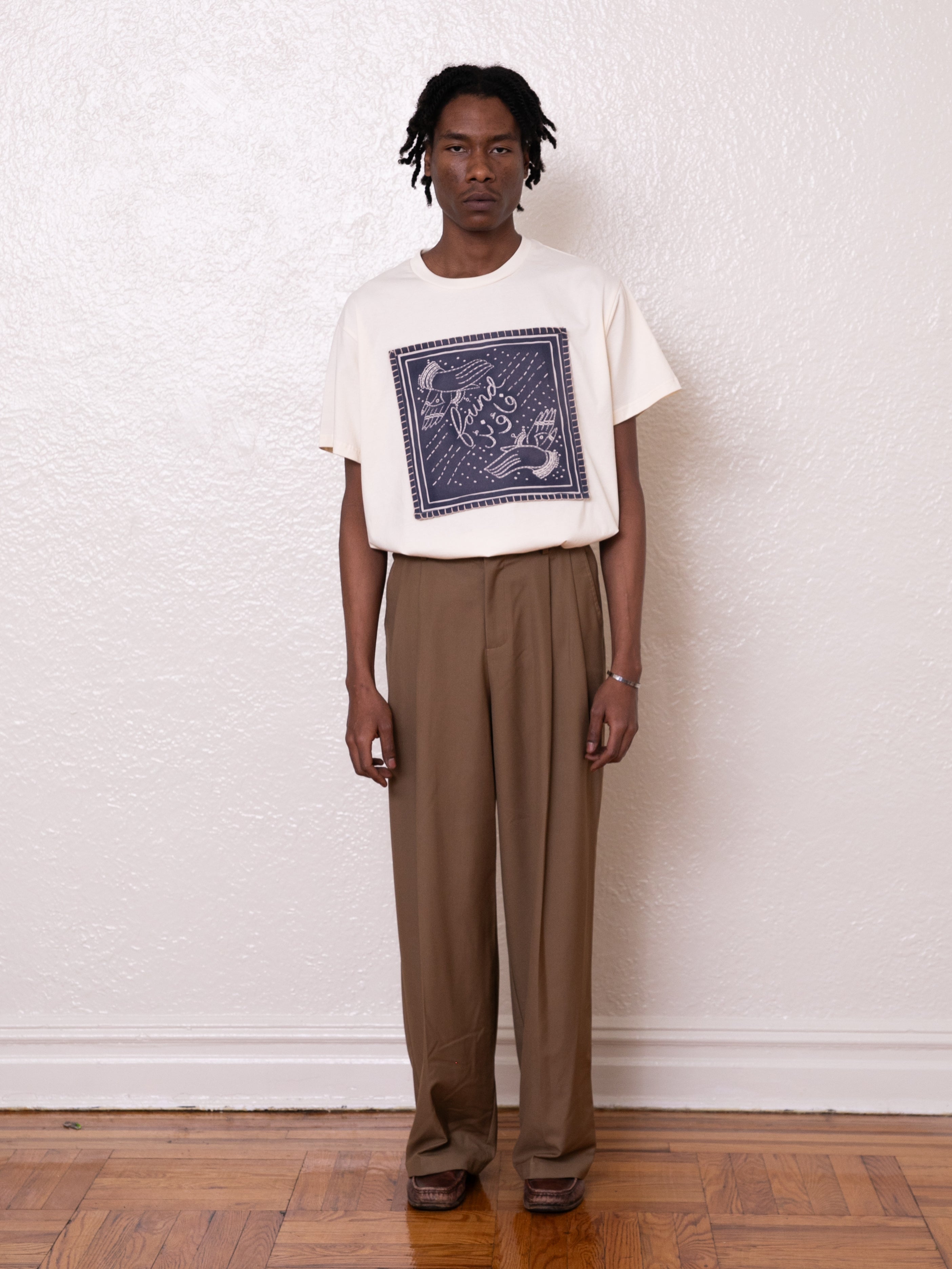 A person stands against a white wall wearing a Hand Art Graphic Tee from FOUND and brown cotton twill trousers, complemented by brown shoes.