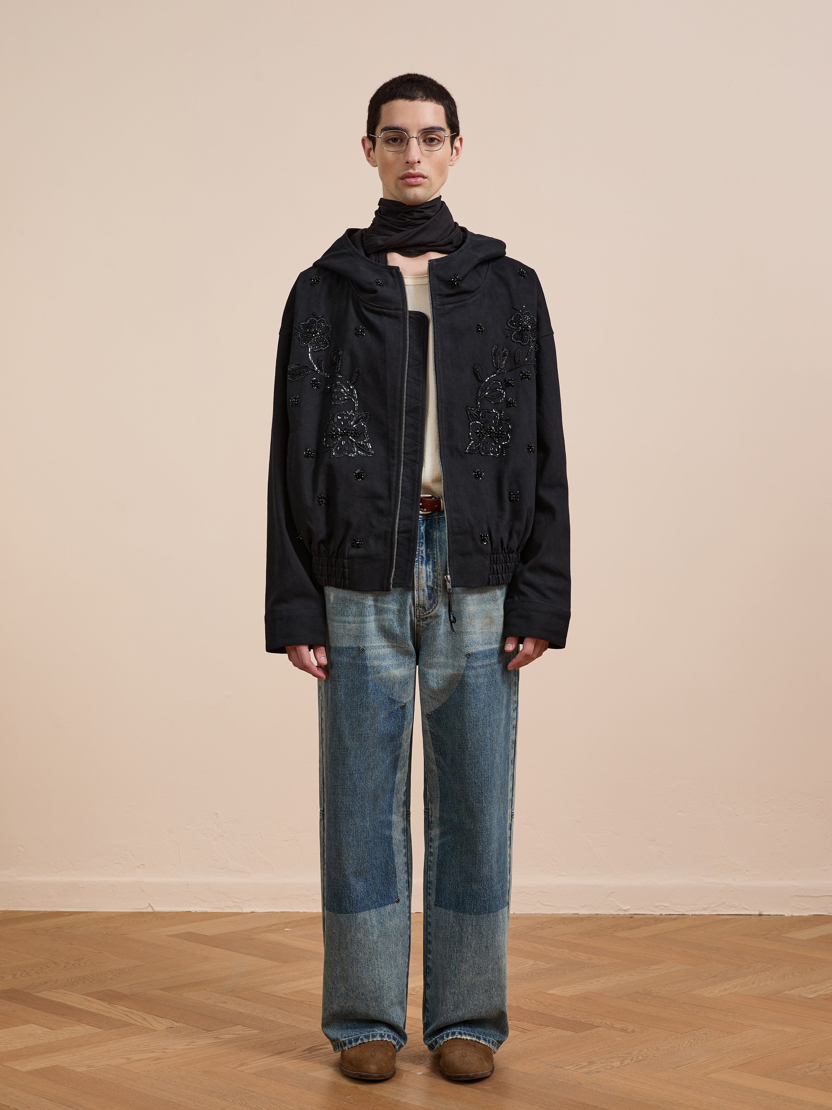 A person in glasses stands against a beige wall wearing the FOUND Hand Embroidered Tonal Anorak Jacket, a scarf, light shirt, wide-leg patchwork cotton canvas jeans, and brown shoes on a wooden floor.