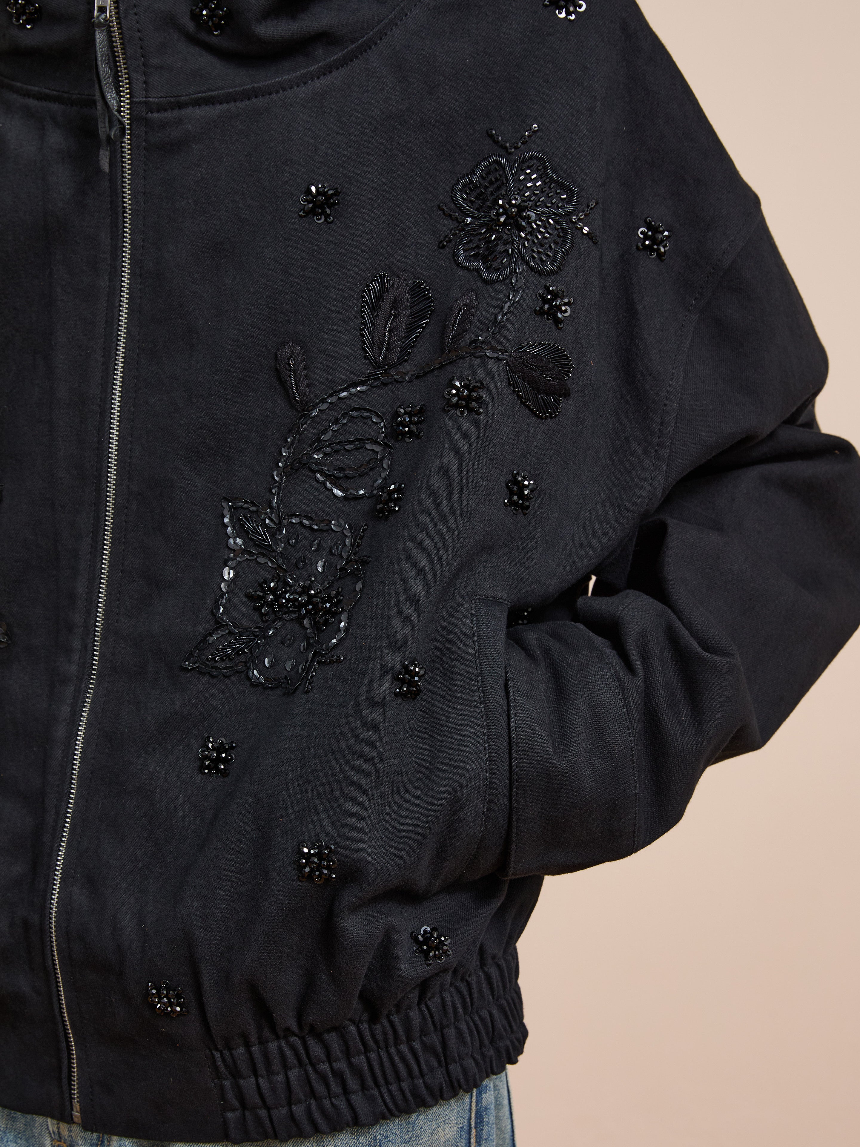 A close-up of FOUND's Hand Embroidered Tonal Anorak Jacket, highlighting intricate black floral embroidery and bead details, with hands casually placed in the side pockets.