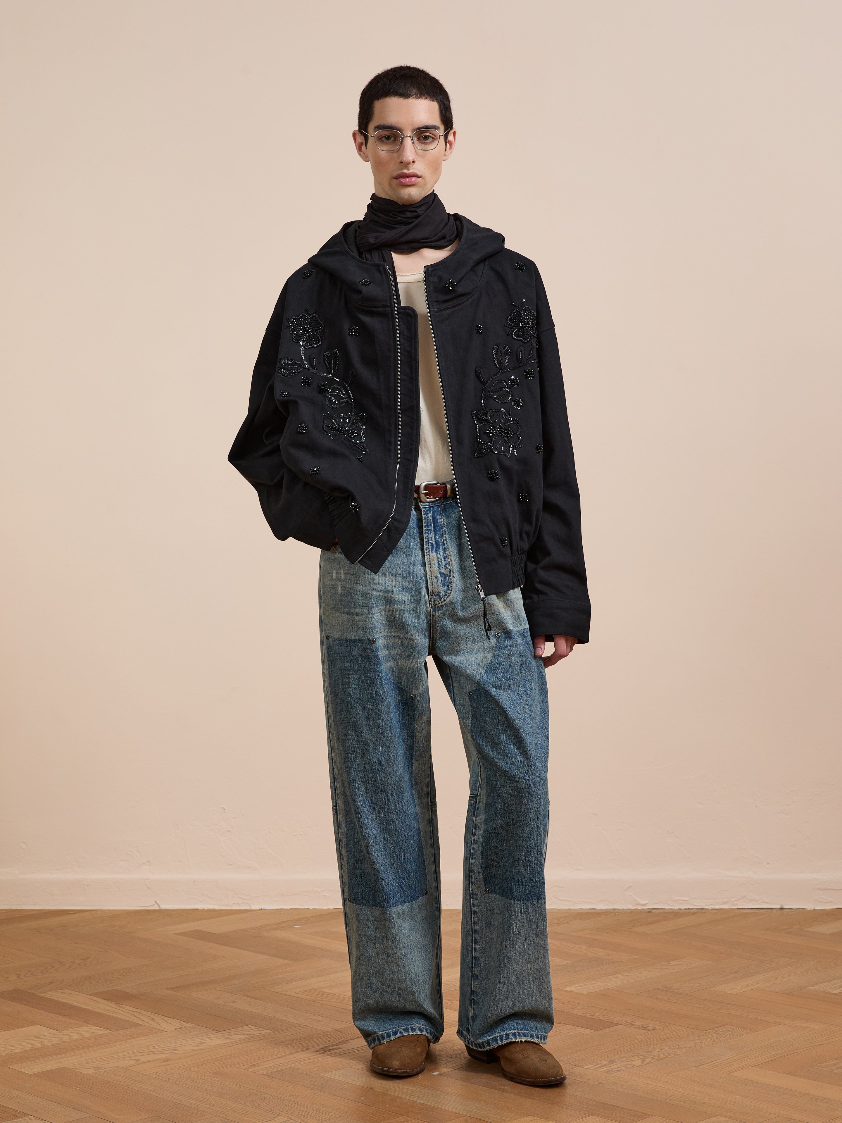 Indoors, a person wears glasses, a black scarf, FOUND's Hand Embroidered Tonal Anorak Jacket, a white shirt, loose patchwork jeans, and brown shoes on a wooden floor with a neutral background.