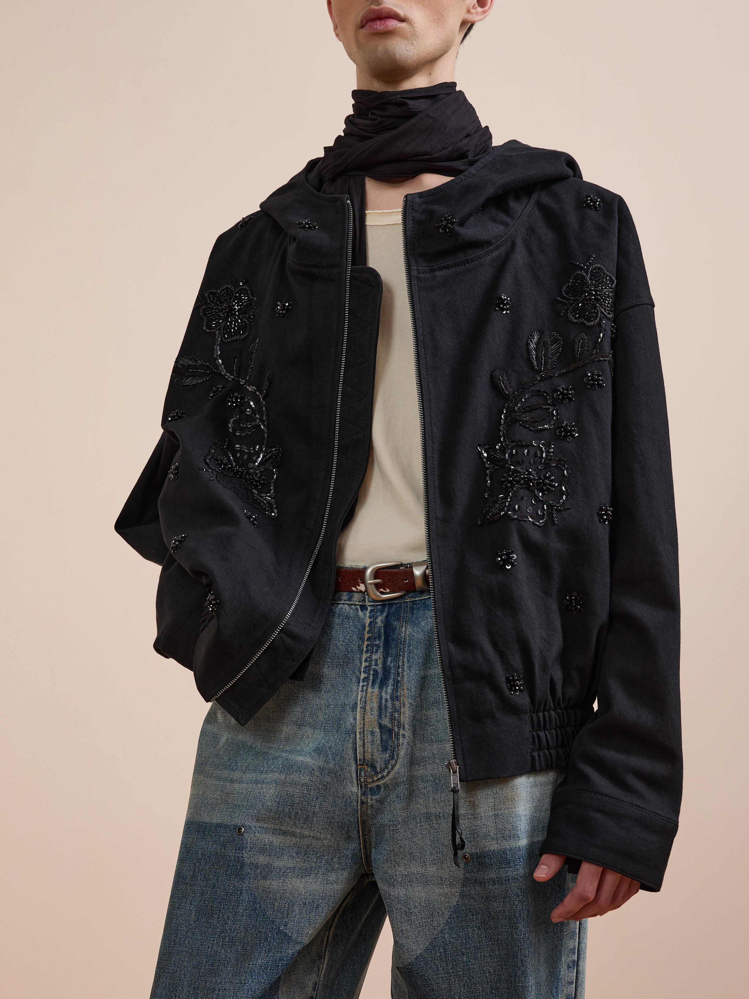 A person wears the FOUND Hand Embroidered Tonal Anorak Jacket over a beige shirt, blue jeans, brown belt, and black scarf, standing against a plain beige background.