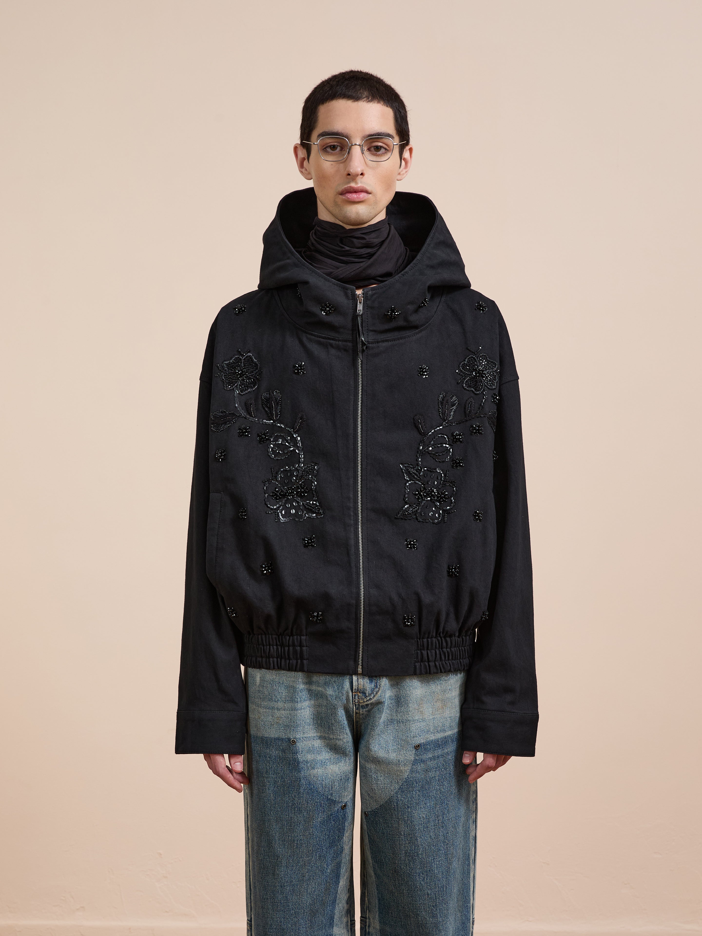 A person in glasses and blue jeans wears the FOUND Hand Embroidered Tonal Anorak Jacket, featuring intricate embroidery, while standing against a plain beige background.