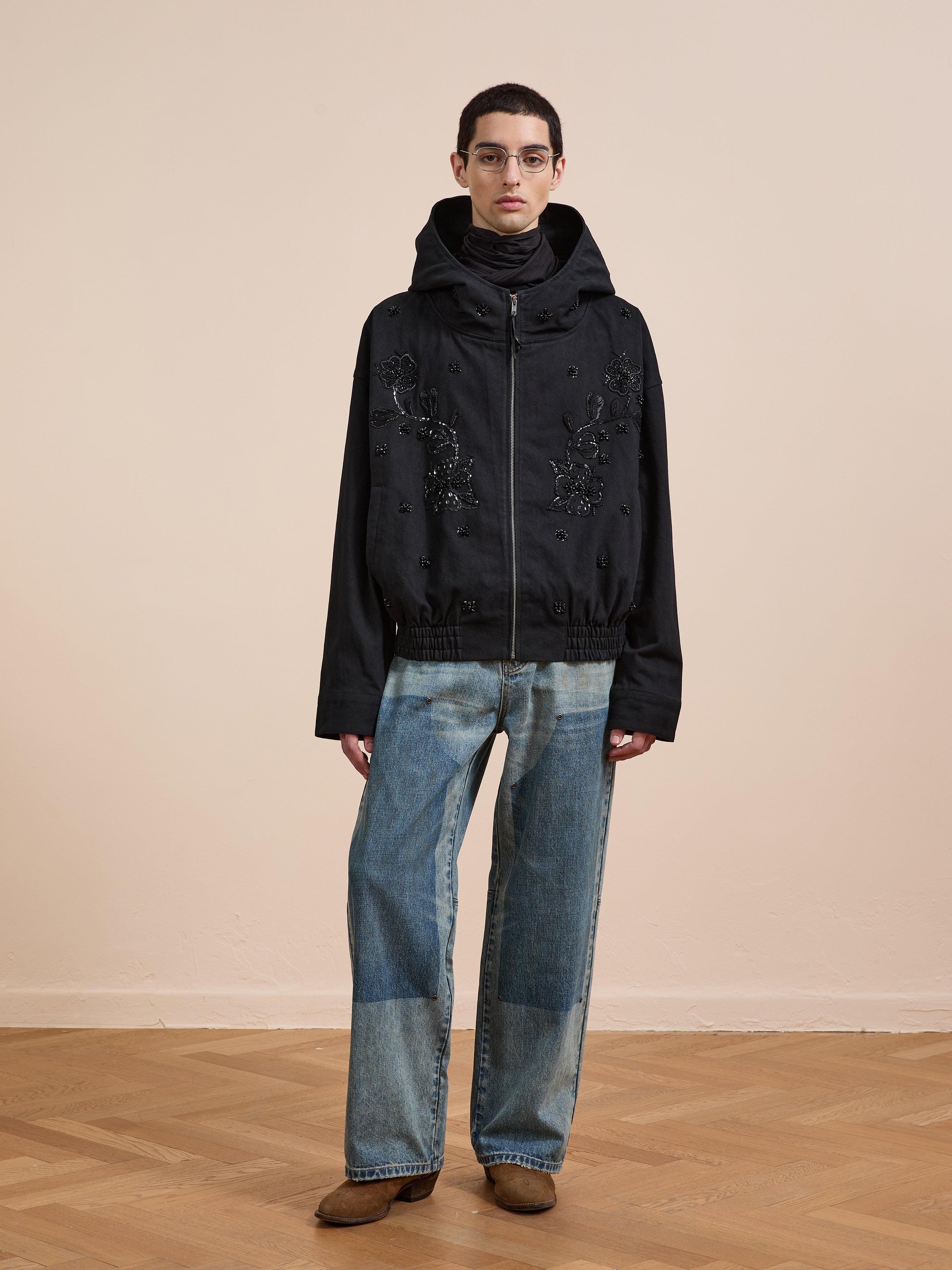 A person stands indoors against a beige wall and wooden floor, wearing a black FOUND Hand Embroidered Tonal Anorak Jacket with embellishments, blue wide-leg patchwork jeans, and brown shoes.