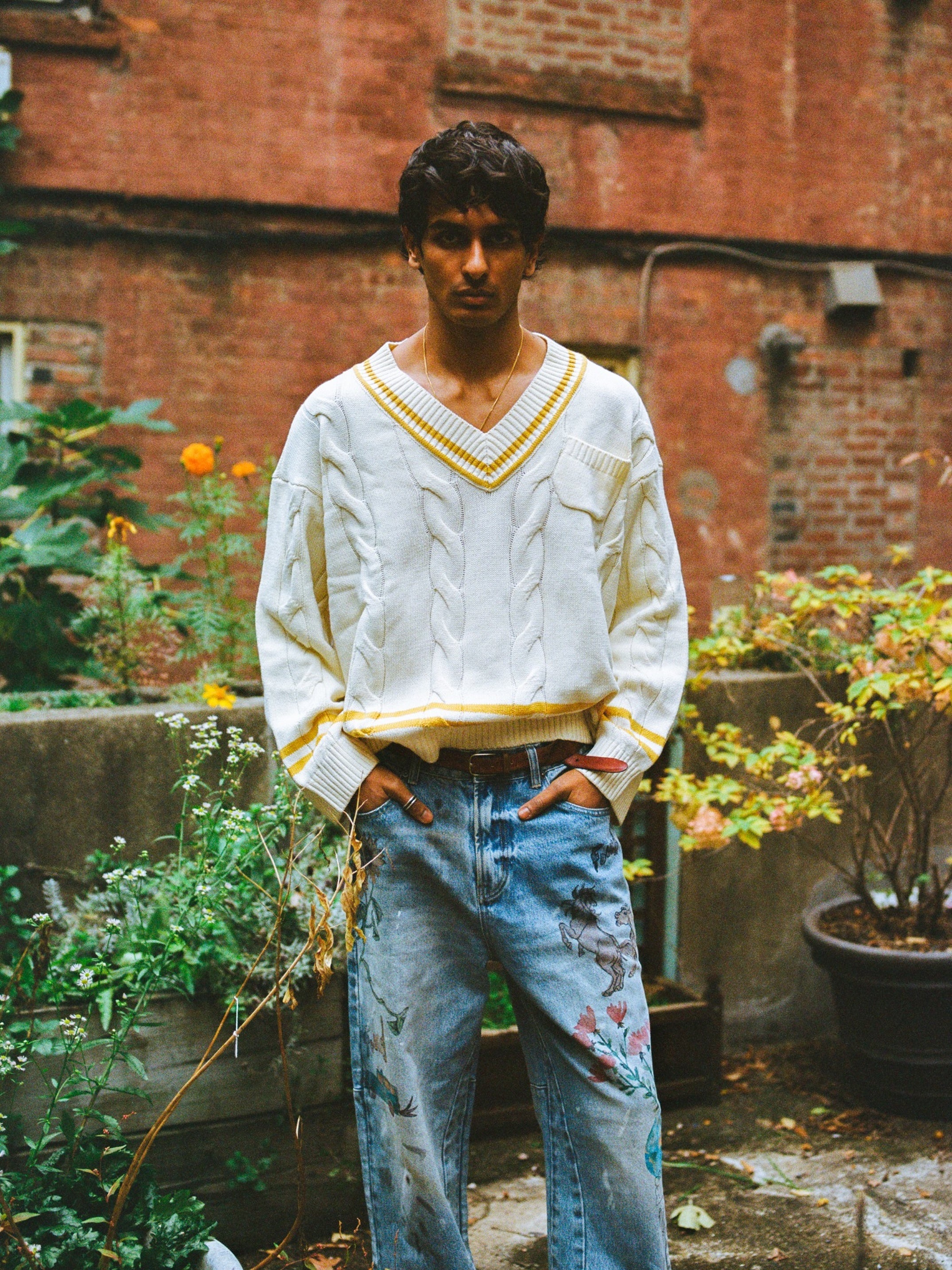 A man stands outdoors wearing a white cable-knit sweater with yellow trim and FOUND Hand Art Jeans—light blue denim featuring hand-drawn floral prints.