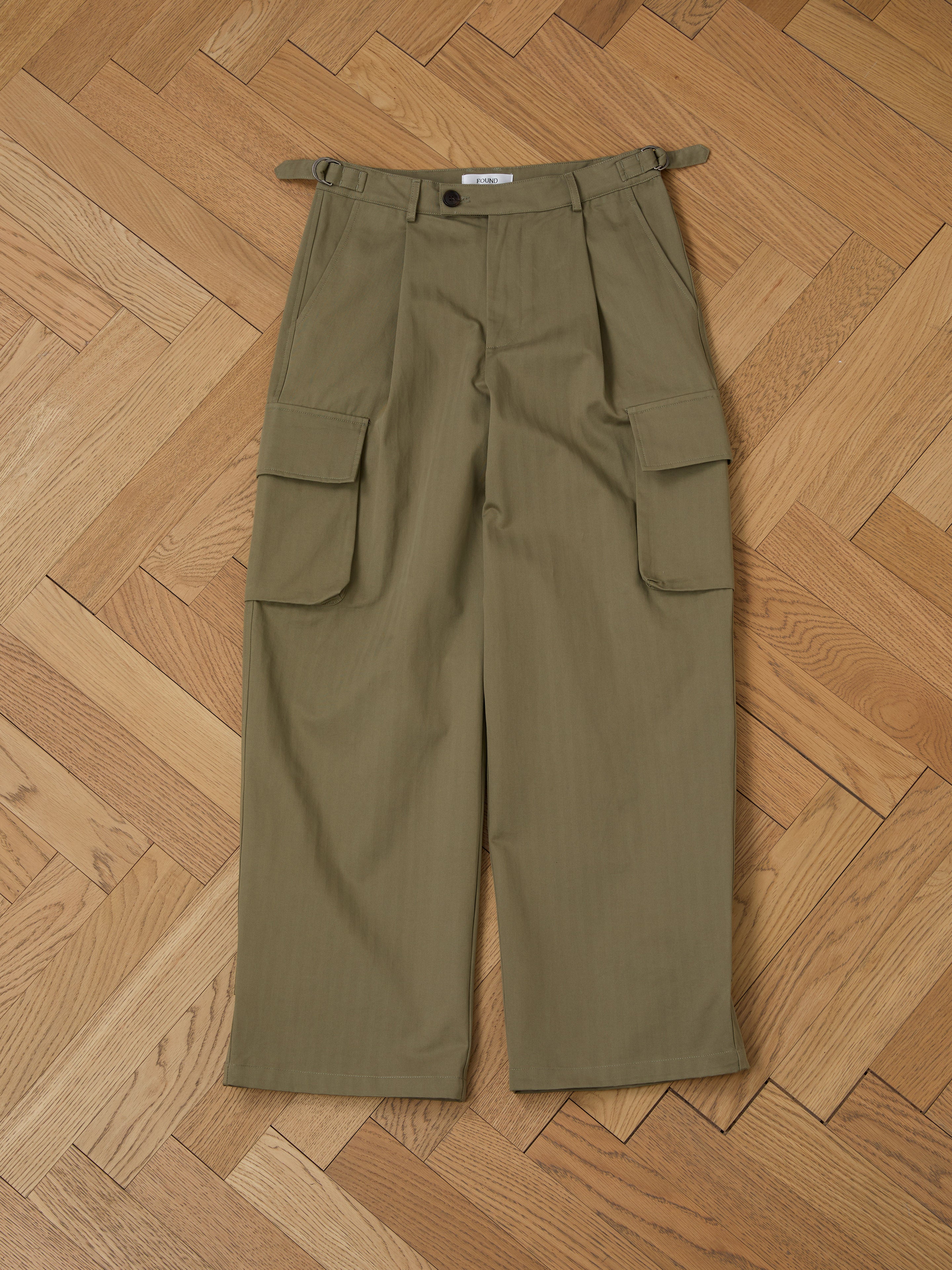 The olive Hava Herringbone Pleated Cargo Trousers by FOUND, made from a wool-blend fabric and featuring side pockets, are displayed flat on a herringbone wood-patterned floor.