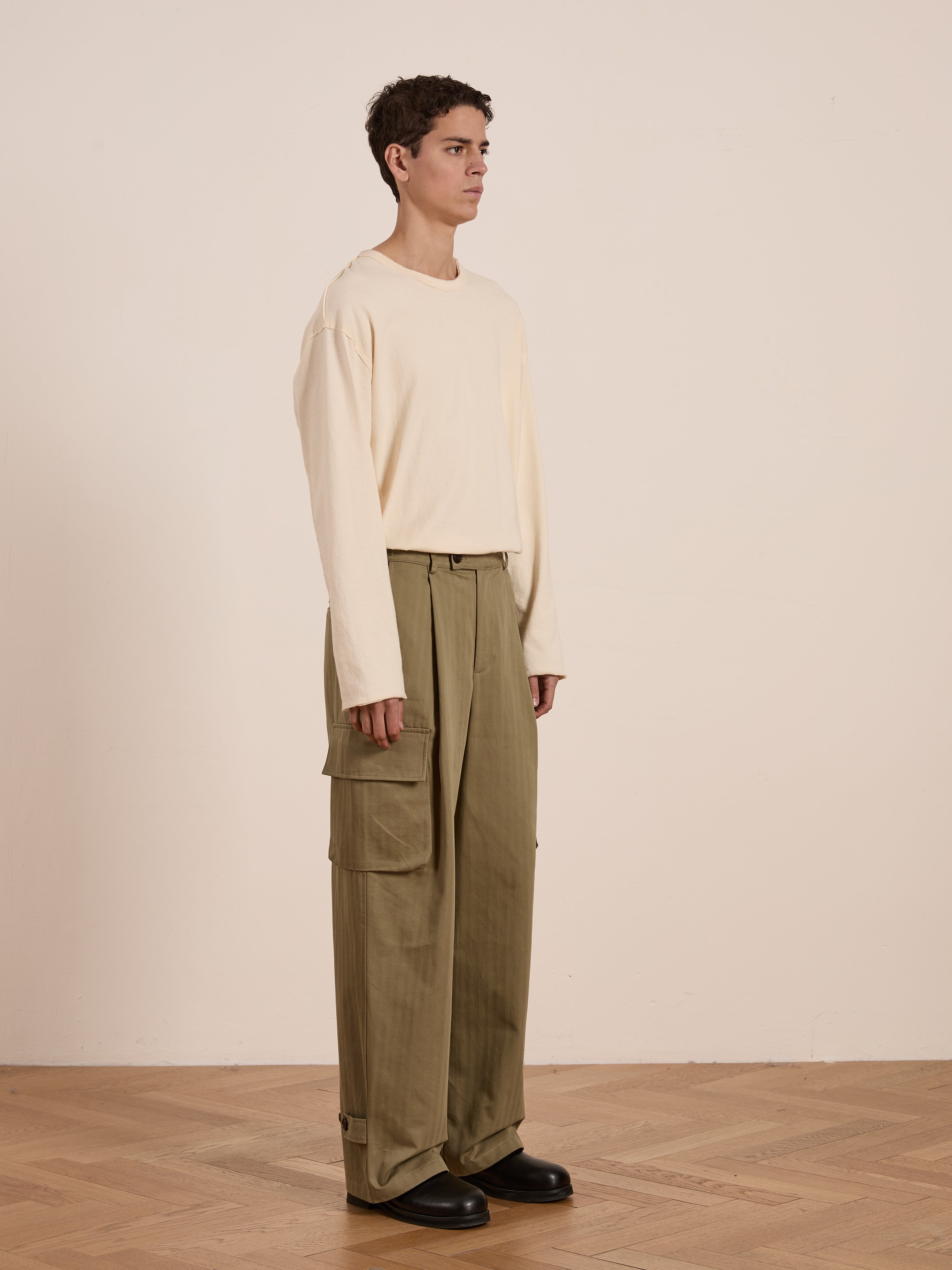 Against an off-white wall and wooden floor, the short-haired individual wears a beige long-sleeve shirt, black shoes, and FOUND's Hava Herringbone Pleated Cargo Trousers in olive green, epitomizing utility fashion.