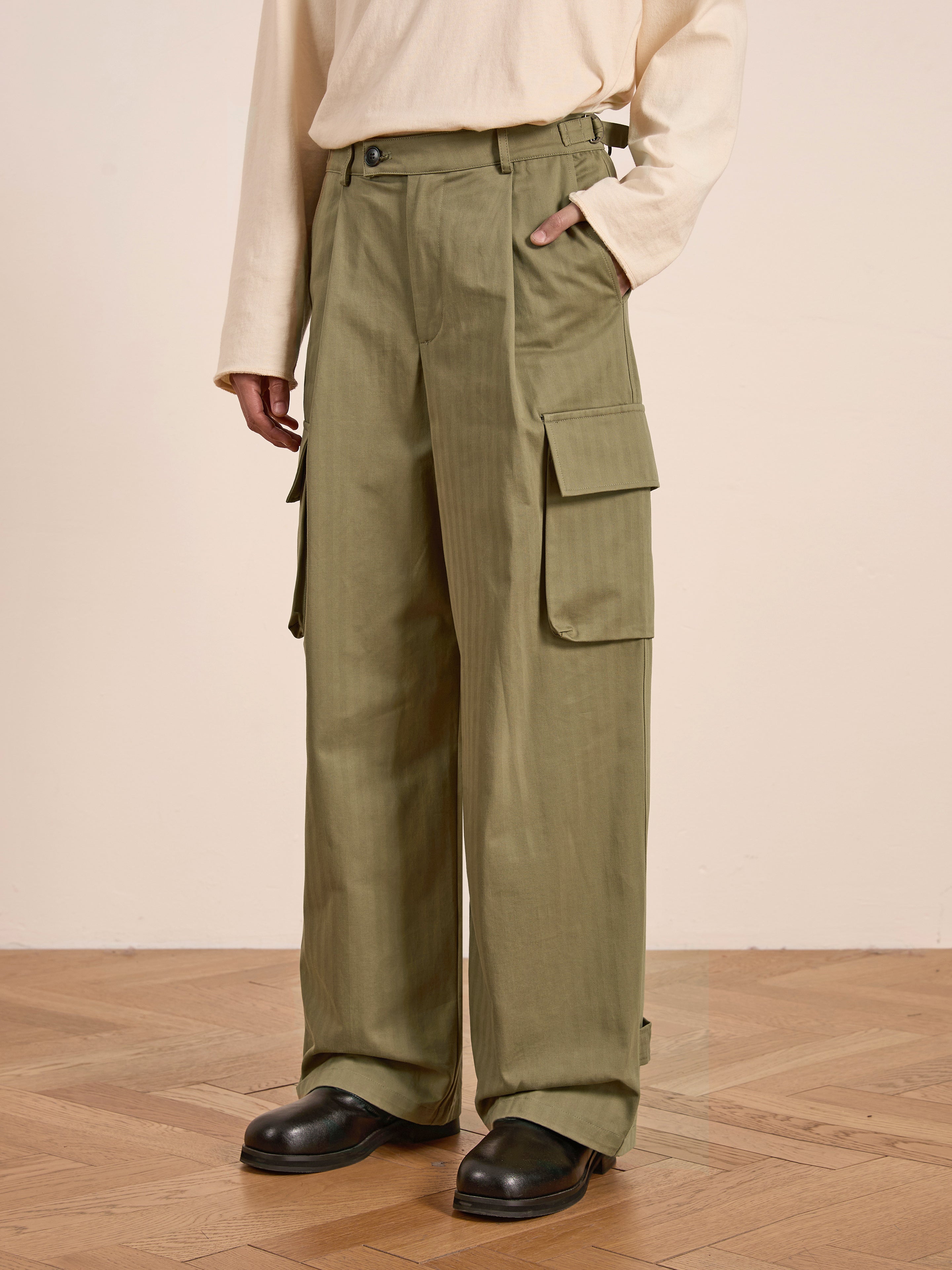 The ensemble features the Hava Herringbone Pleated Cargo Trousers by FOUND in olive green, paired with a beige long-sleeve top and black shoes on wooden flooring.