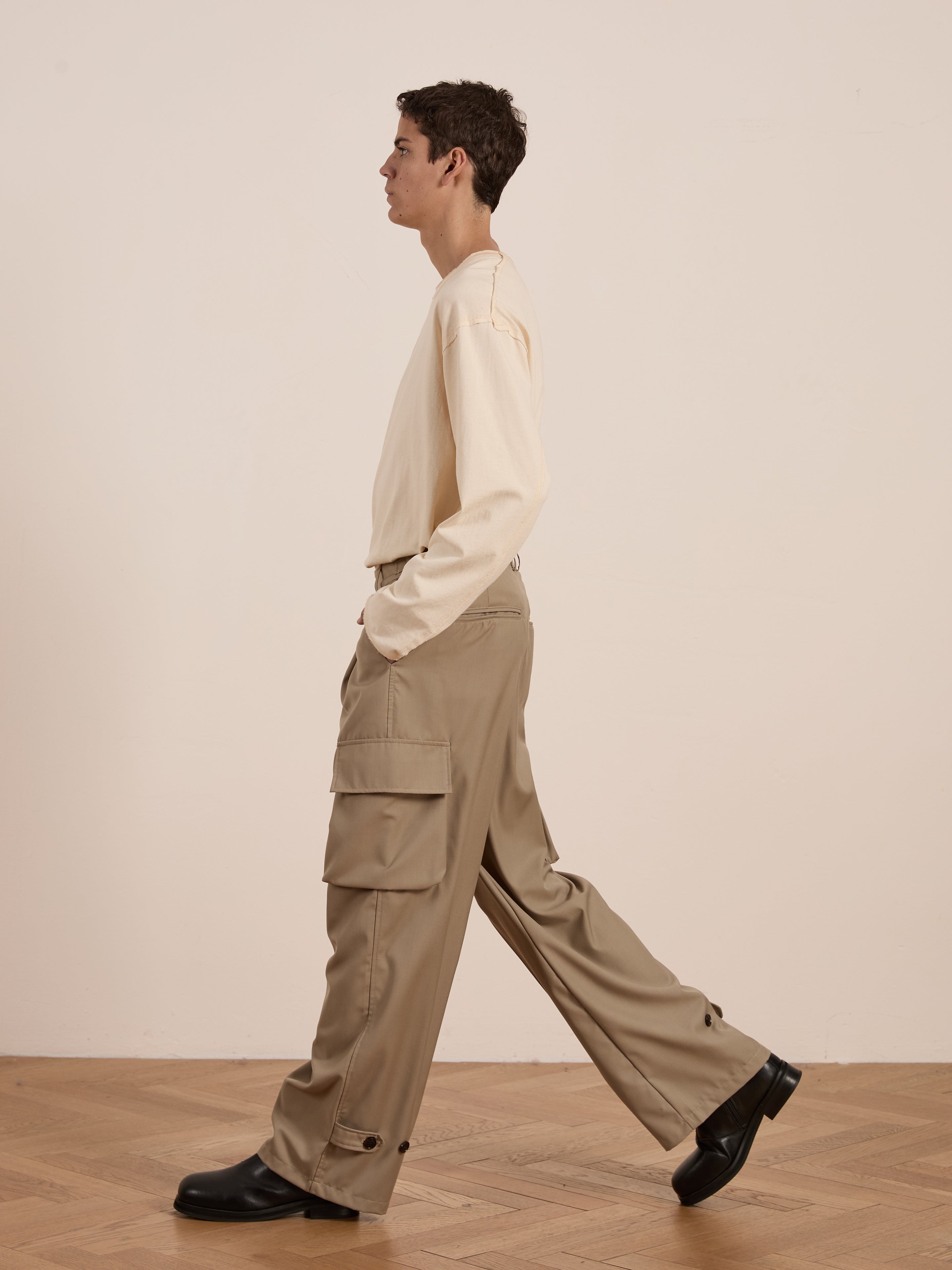 Sporting the utilitarian style, someone in a cream shirt and FOUND's beige Hava Pleated Cargo Trousers strolls on a wooden floor.