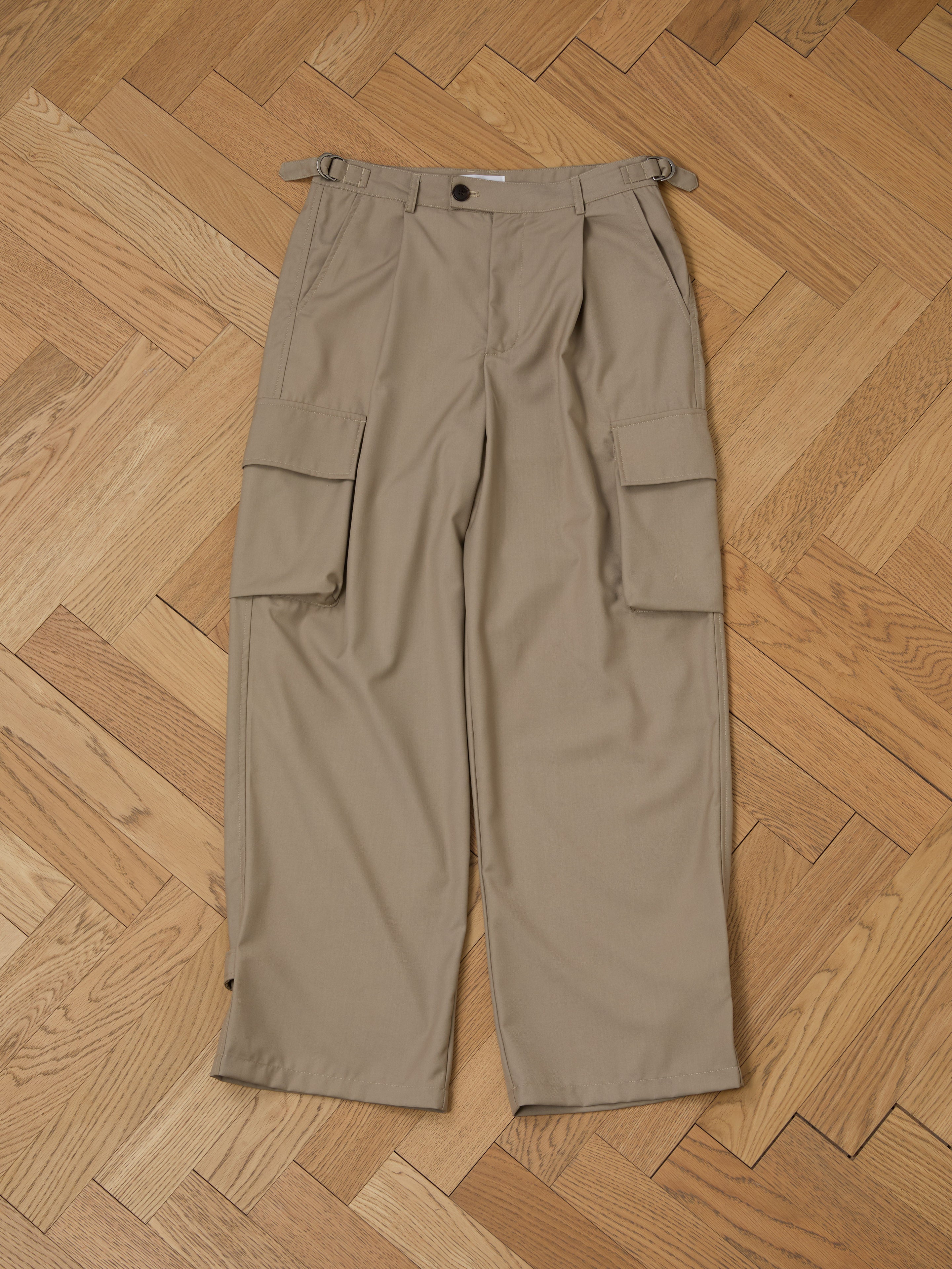 FOUND's Hava Pleated Cargo Trousers, made from durable cotton twill, lie flat on a herringbone wooden floor, showing off their utilitarian style and multiple pockets.