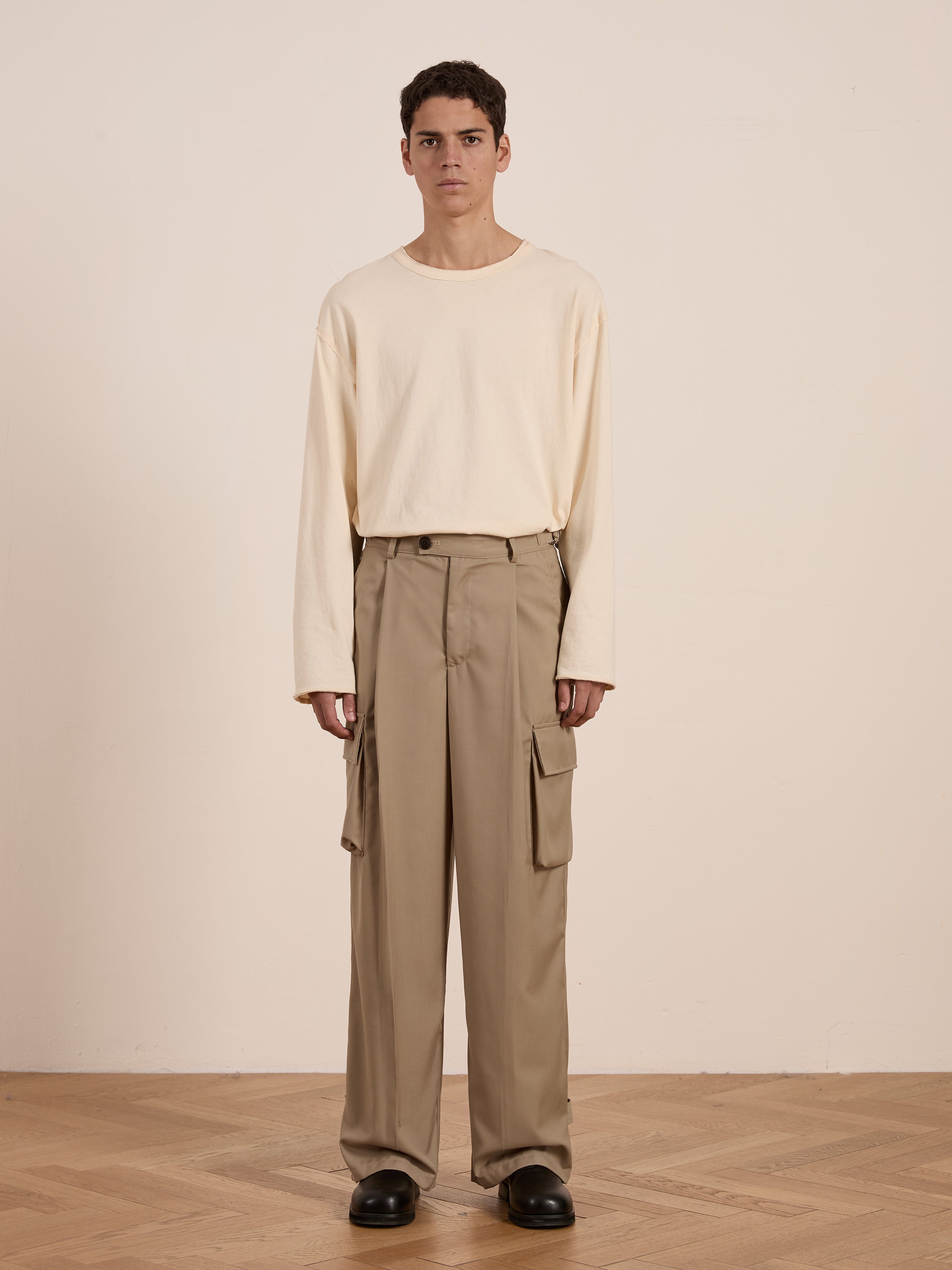 Indoors, a person stands on a wooden floor wearing cream long-sleeve shirt, black shoes, and FOUND's Hava Pleated Cargo Trousers in a utilitarian khaki style.