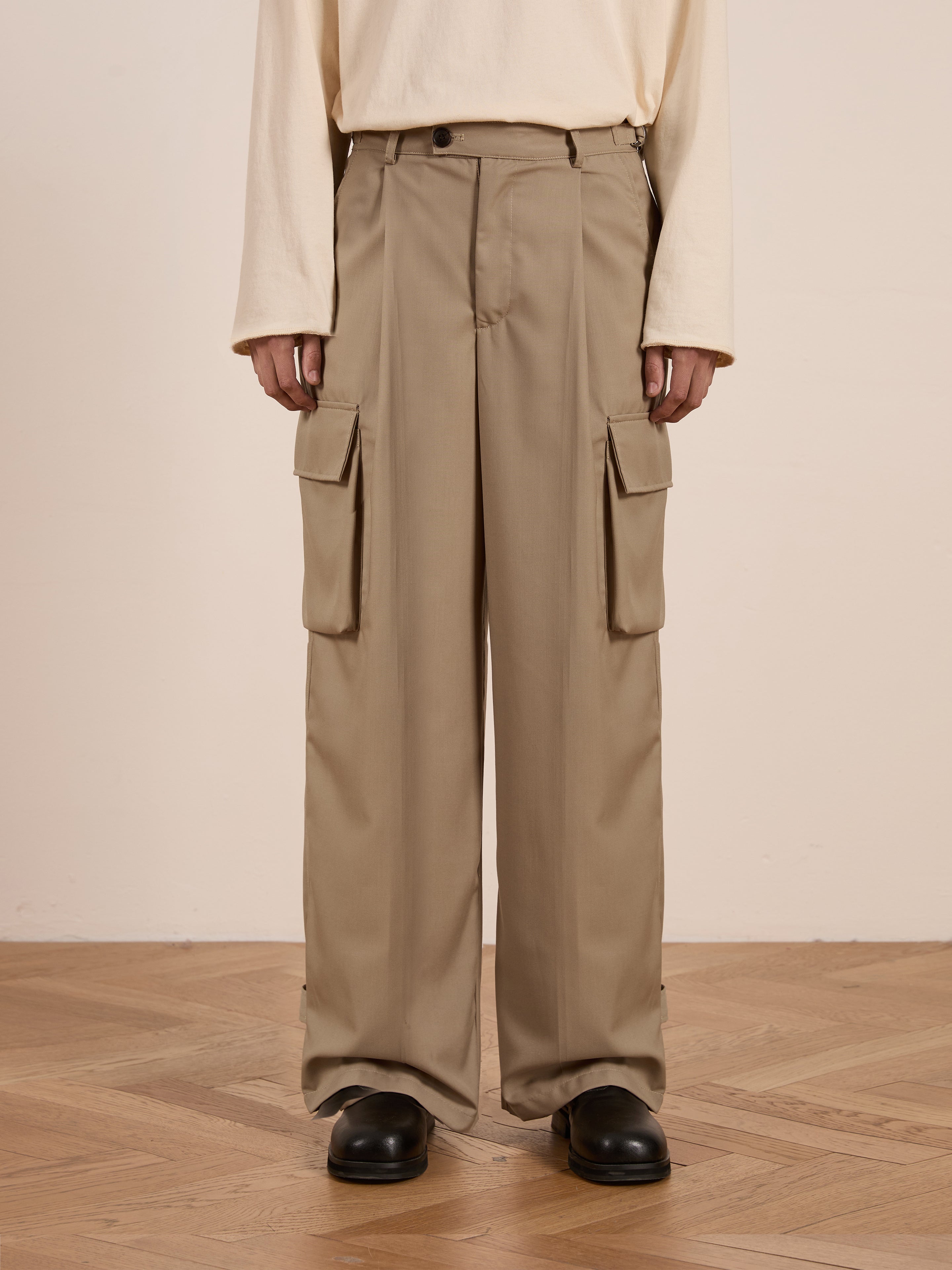 Wearing FOUND's Hava Pleated Cargo Trousers in beige with large side pockets, paired with a cream long-sleeve shirt, this person stylishly showcases unisex fashion while standing on a wooden floor.