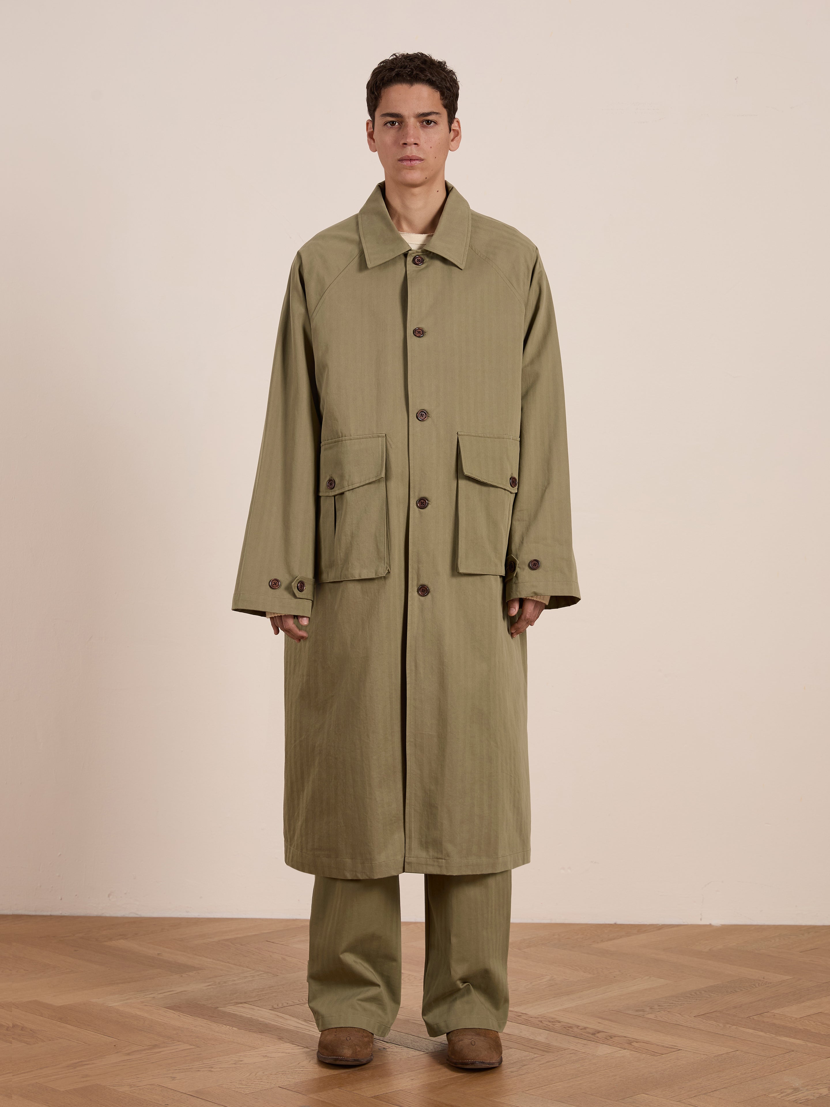 A person indoors wears an olive green Herringbone Twill Trench Coat by FOUND, featuring utility pockets. Matching pants and brown shoes complete the ensemble against a neutral background.