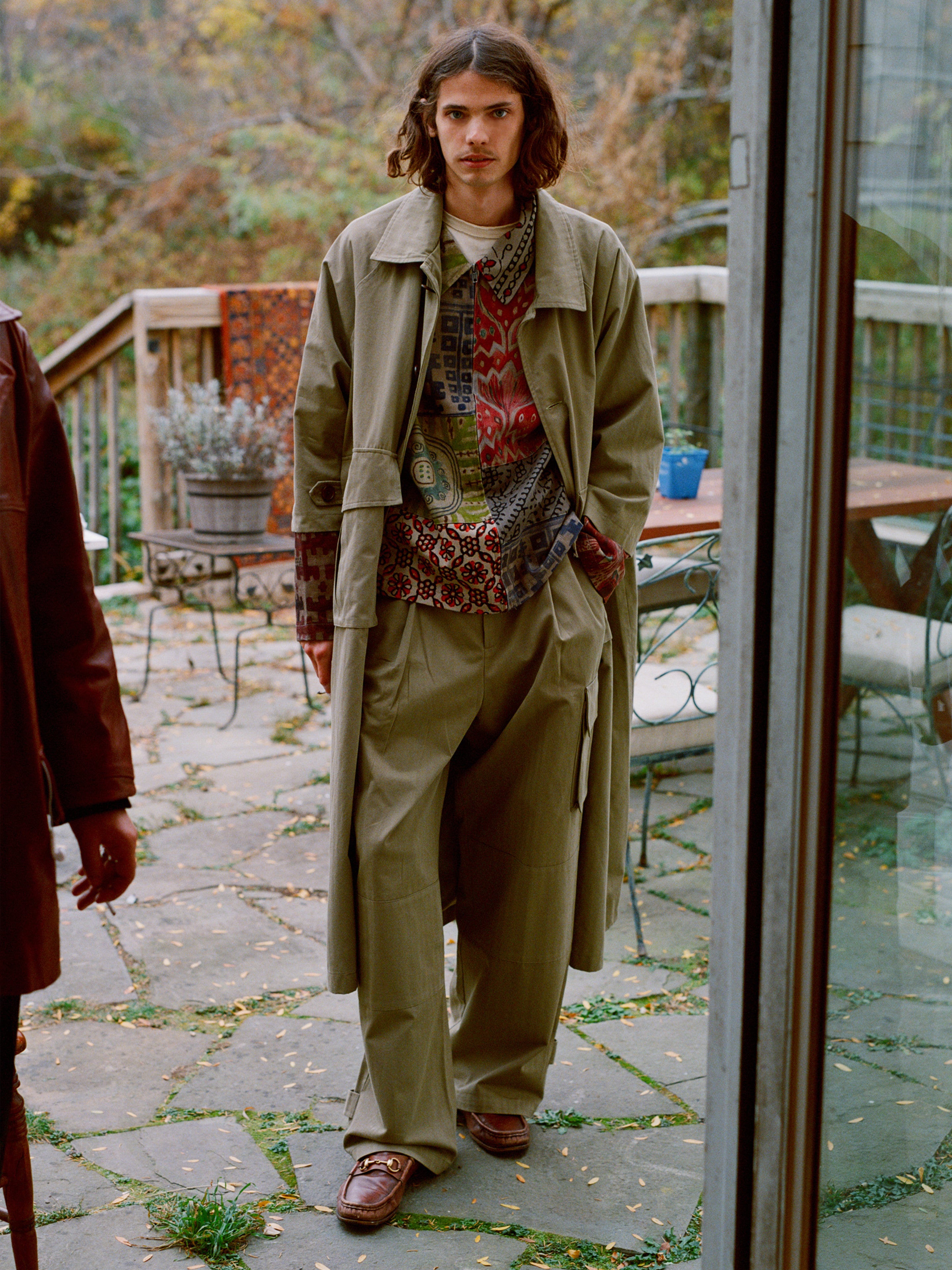 On a patio framed by autumn foliage, someone with long hair wears the FOUND Herringbone Twill Trench Coat and relaxed wide pants, along with brown shoes. Their hand casually rests in a utility pocket.