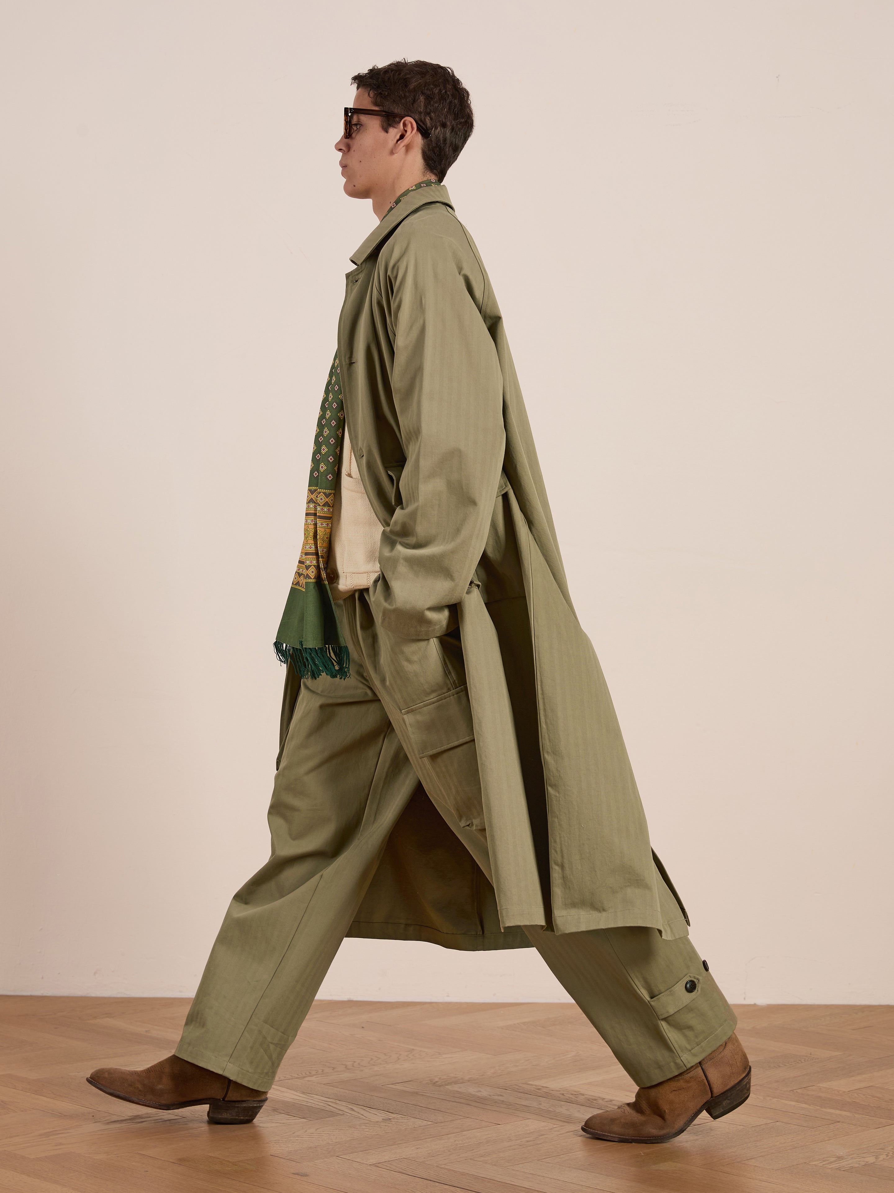 A person confidently walks in profile, wearing FOUND's Herringbone Twill Trench Coat with utility pockets, coordinating pants, sunglasses, brown boots, and a patterned green scarf.