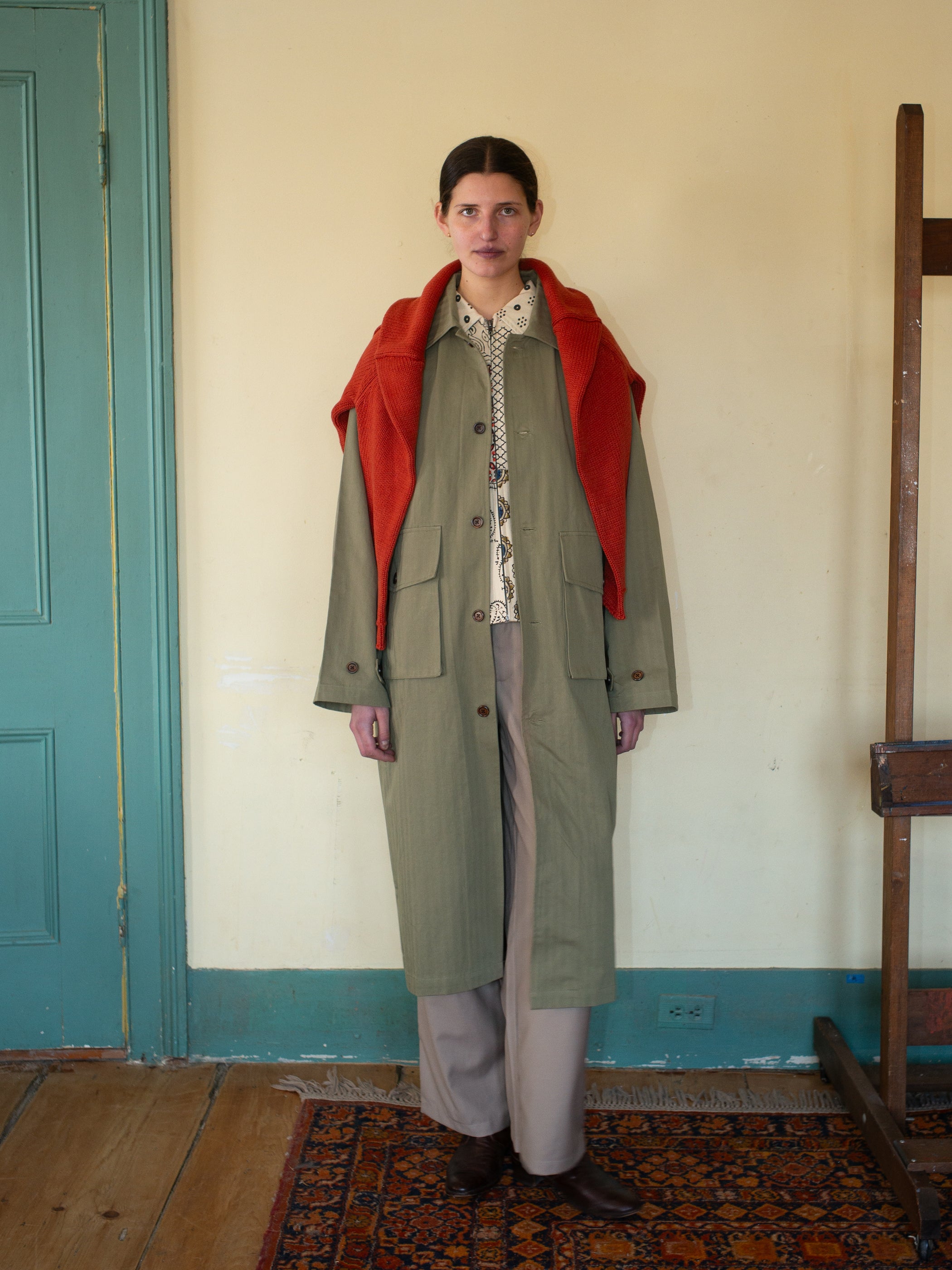 In a room, someone wears FOUND's long green Herringbone Twill Trench Coat over a patterned shirt and pants, with an orange sweater draped over their shoulders. A door and a wooden easel are seen in the background.